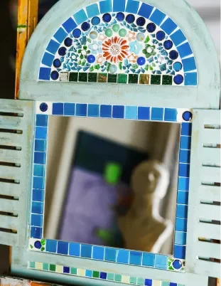 Decorative mirror with a mosaic tile arch and frame featuring blue, green, and orange tiles, reflecting part of a smartphone and a blurred background.