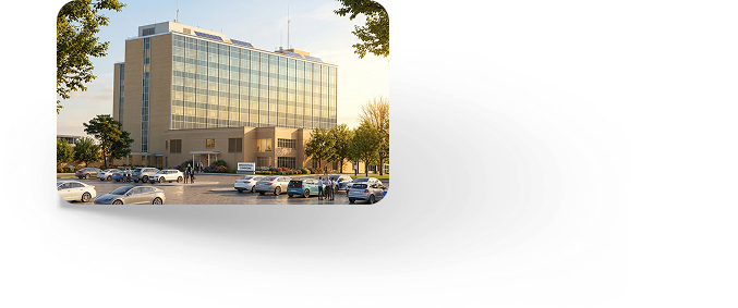 A large modern office building with a glass facade, surrounded by a parking lot with several cars and a few people walking outside, taken during daylight hours.
