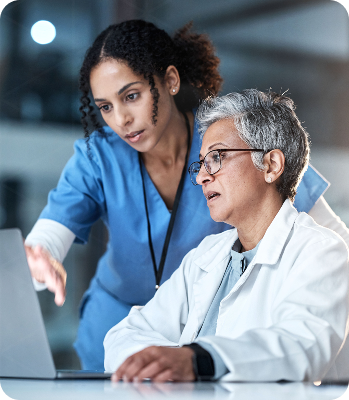 A young woman in blue scrubs and an elderly man in a white lab coat working together on a laptop in a medical setting.