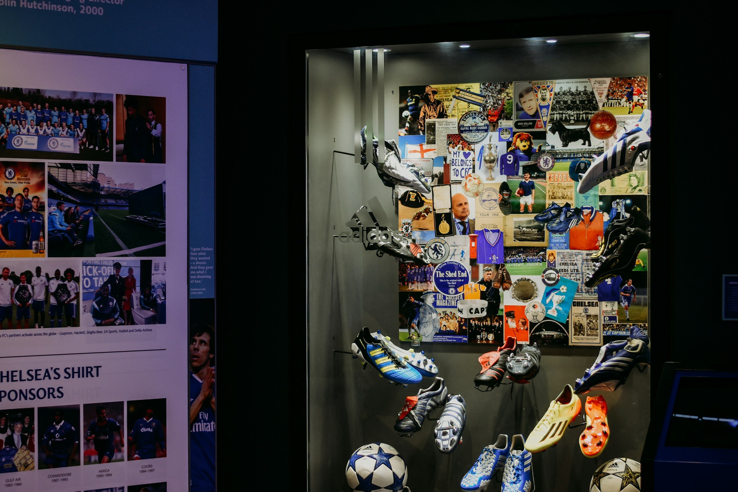 A display case filled with Chelsea football memorabilia including shoes, a ball, photographs, and various keepsakes. To the left, a poster features Chelsea football team photos and related images.