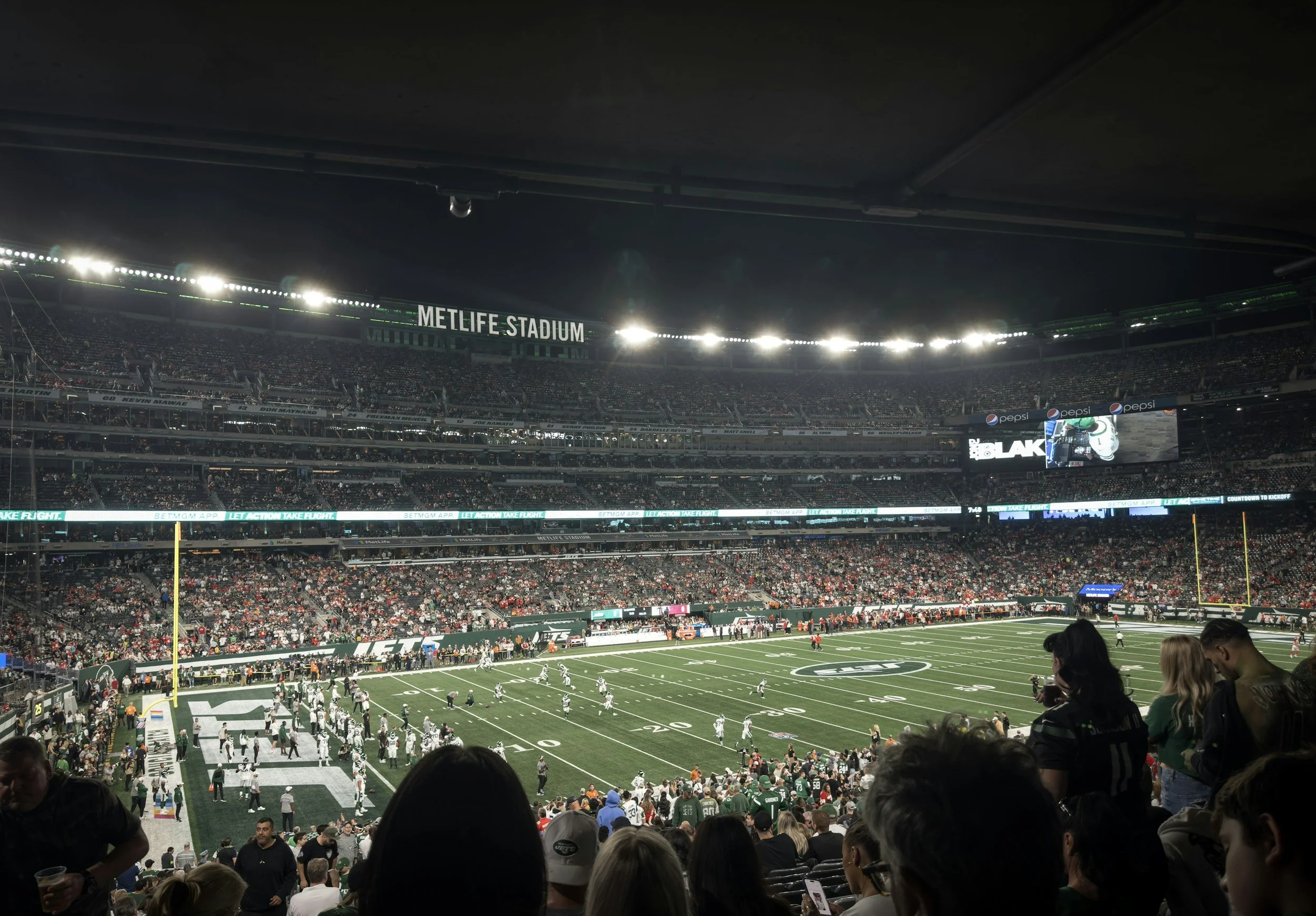 A wide view of MetLife Stadium during a football game, filled with spectators, players on the field, and bright stadium lights illuminating the scene.