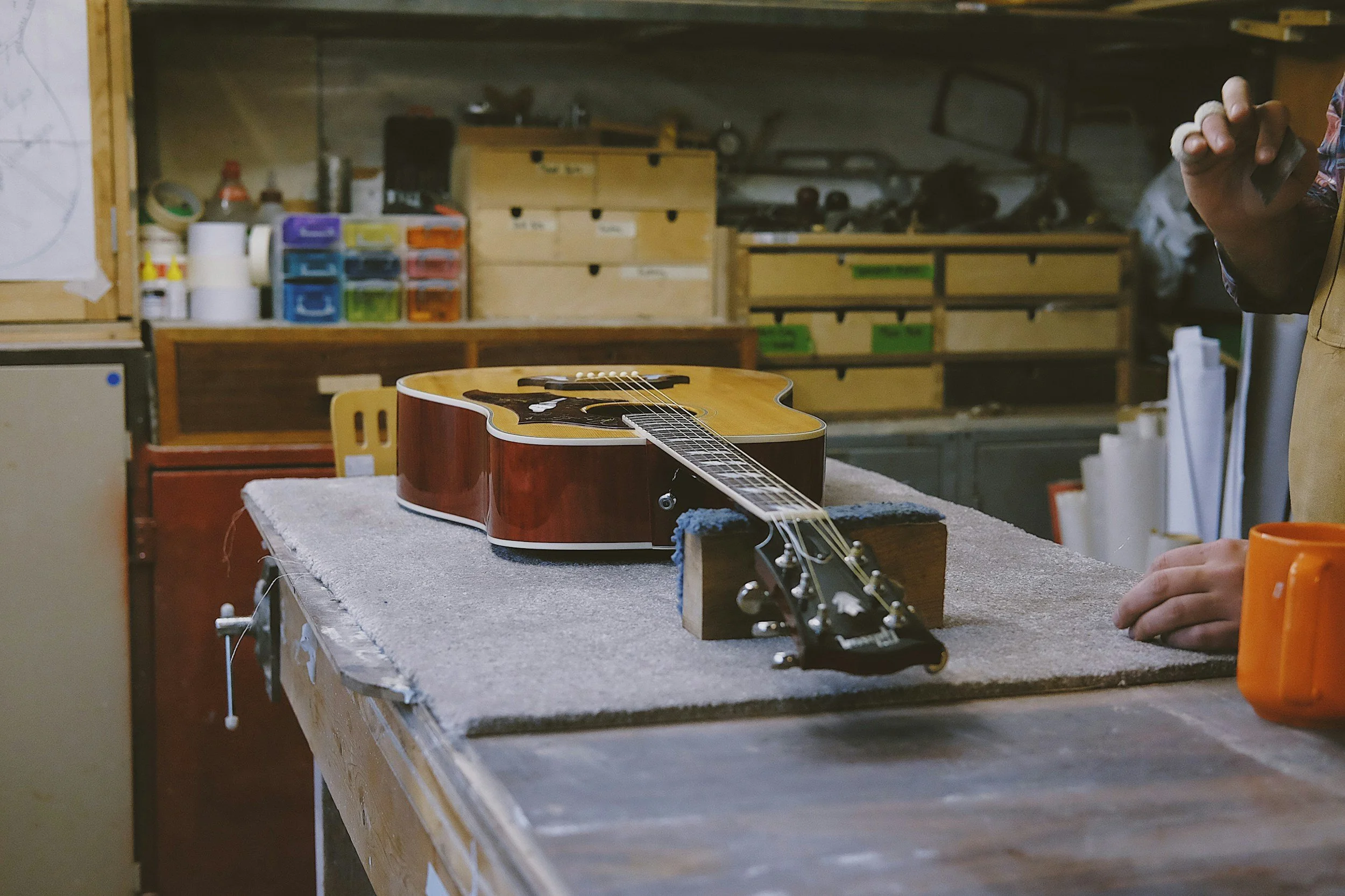 An acoustic guitar placed on a workbench in a woodworking or musical instrument workshop, with a person partially visible on the right side of the image holding a tool or object, and shelves with various supplies and tools in the background.