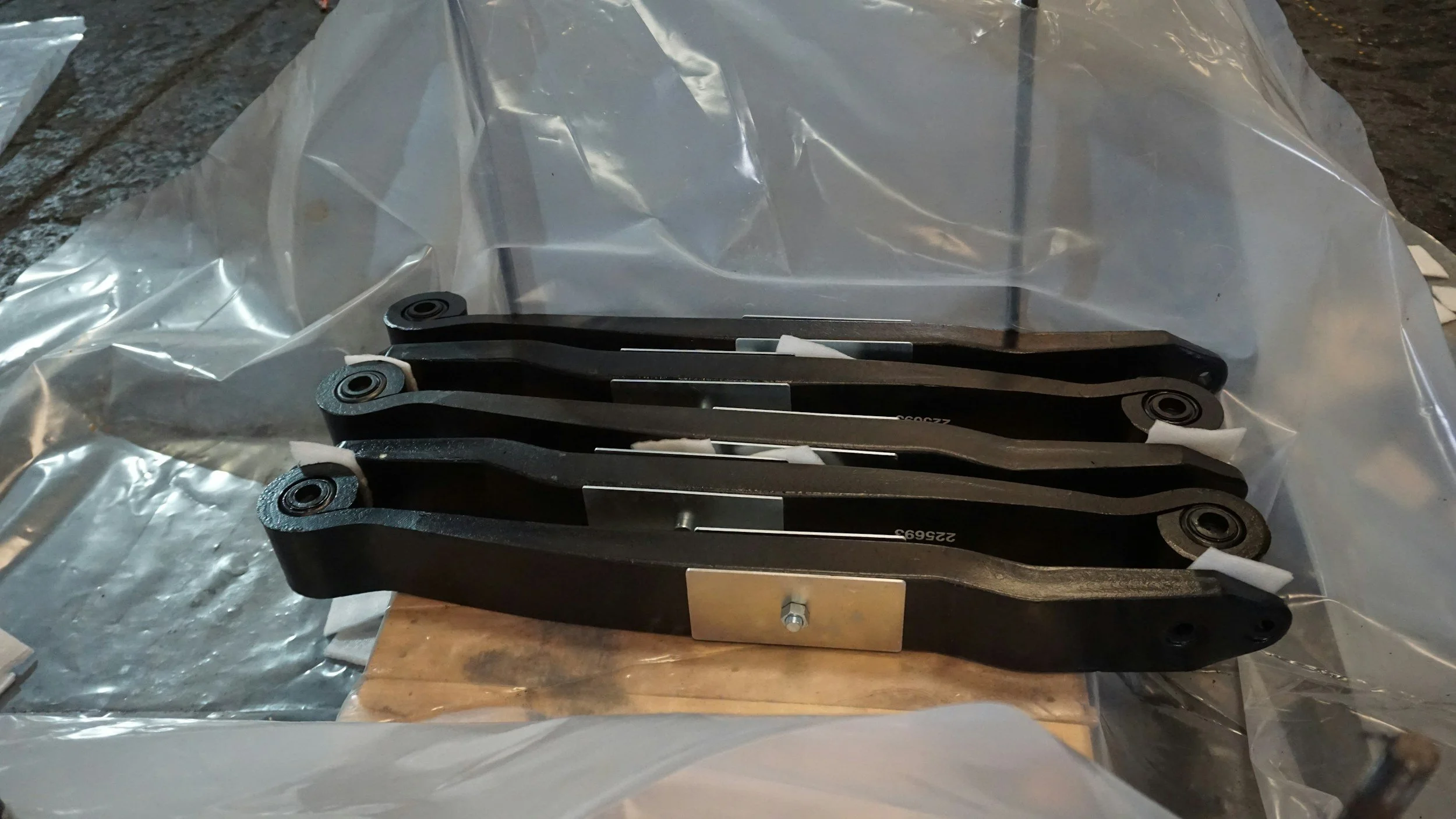 A set of black metal brackets with circular mounting points, placed on a wooden surface and covered with plastic wrappings.