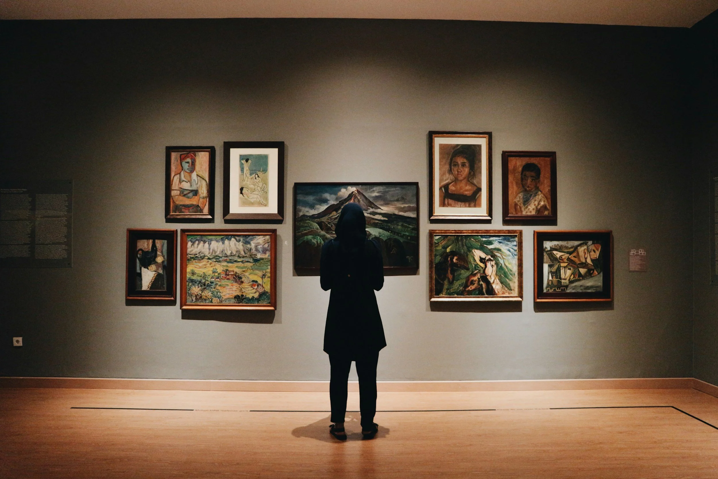 A person standing in front of a wall with multiple framed paintings in an art gallery.