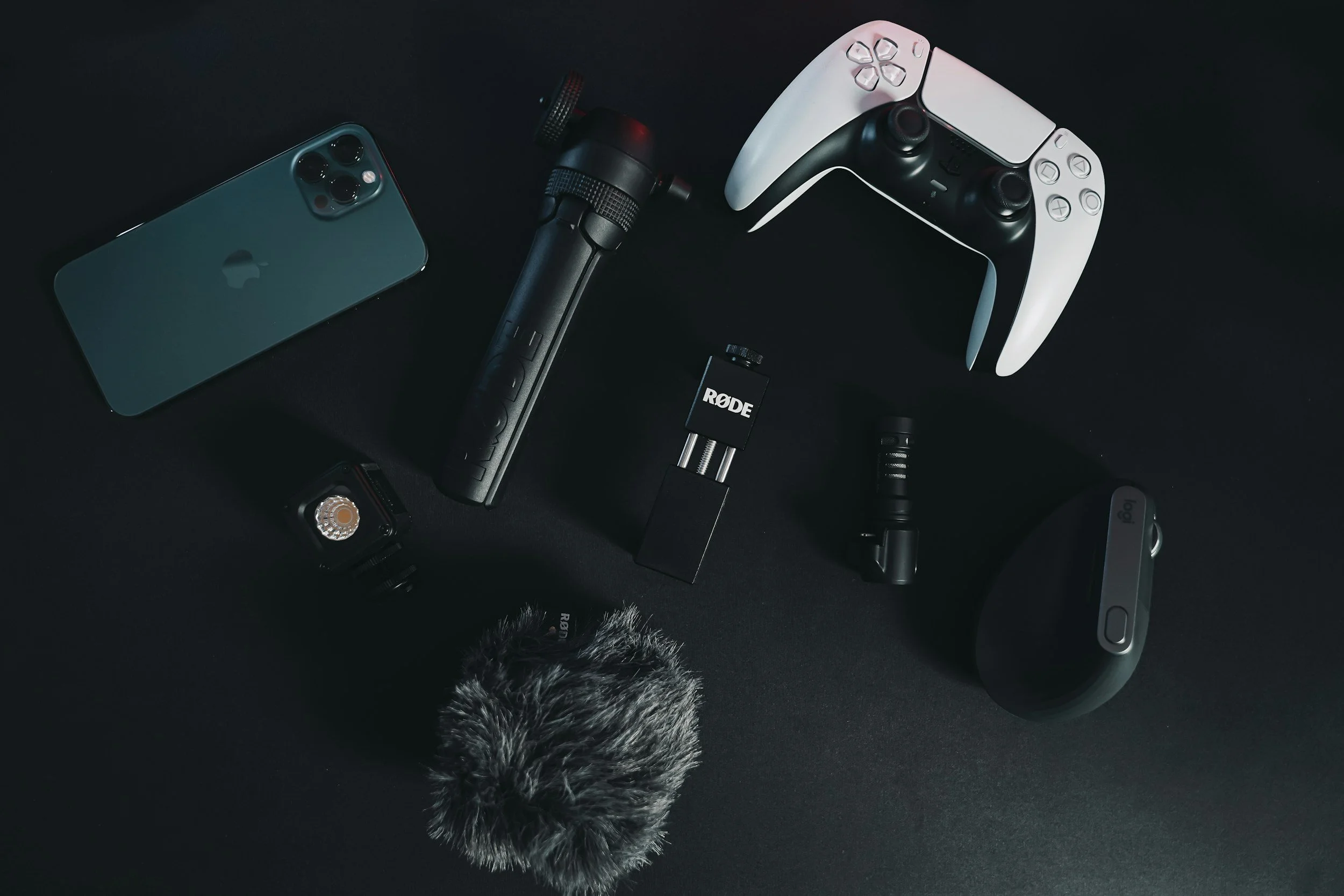 A collection of electronic and audio devices on a black surface, including a green smartphone, a microphone, a camera lens, a portable microphone receiver, and a graphic controller.