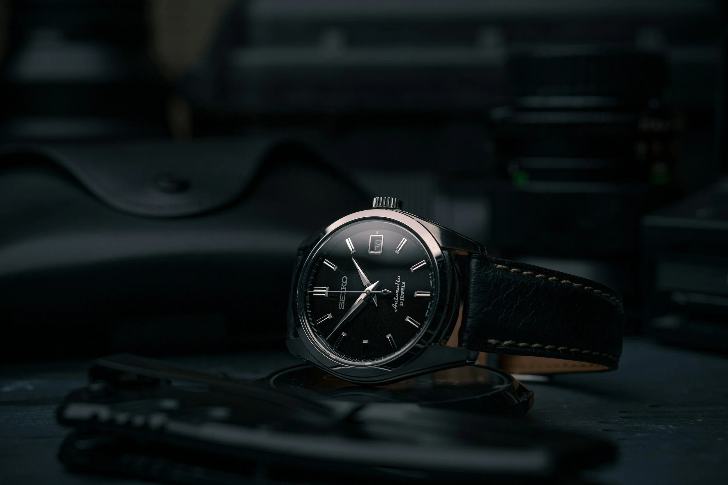 A wristwatch with a black dial, silver case, and black strap is resting on a reflective surface in a dimly lit environment.