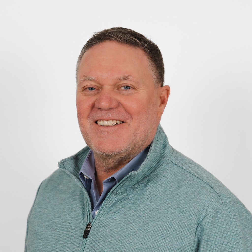 Portrait of a middle-aged man with short brown hair, wearing a light teal zip-up sweater over a collared shirt, smiling against a plain white background.