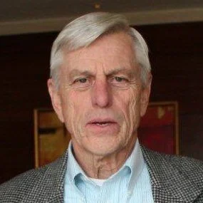 An elderly man with white hair wearing a light blue shirt and a checkered blazer, standing indoors.