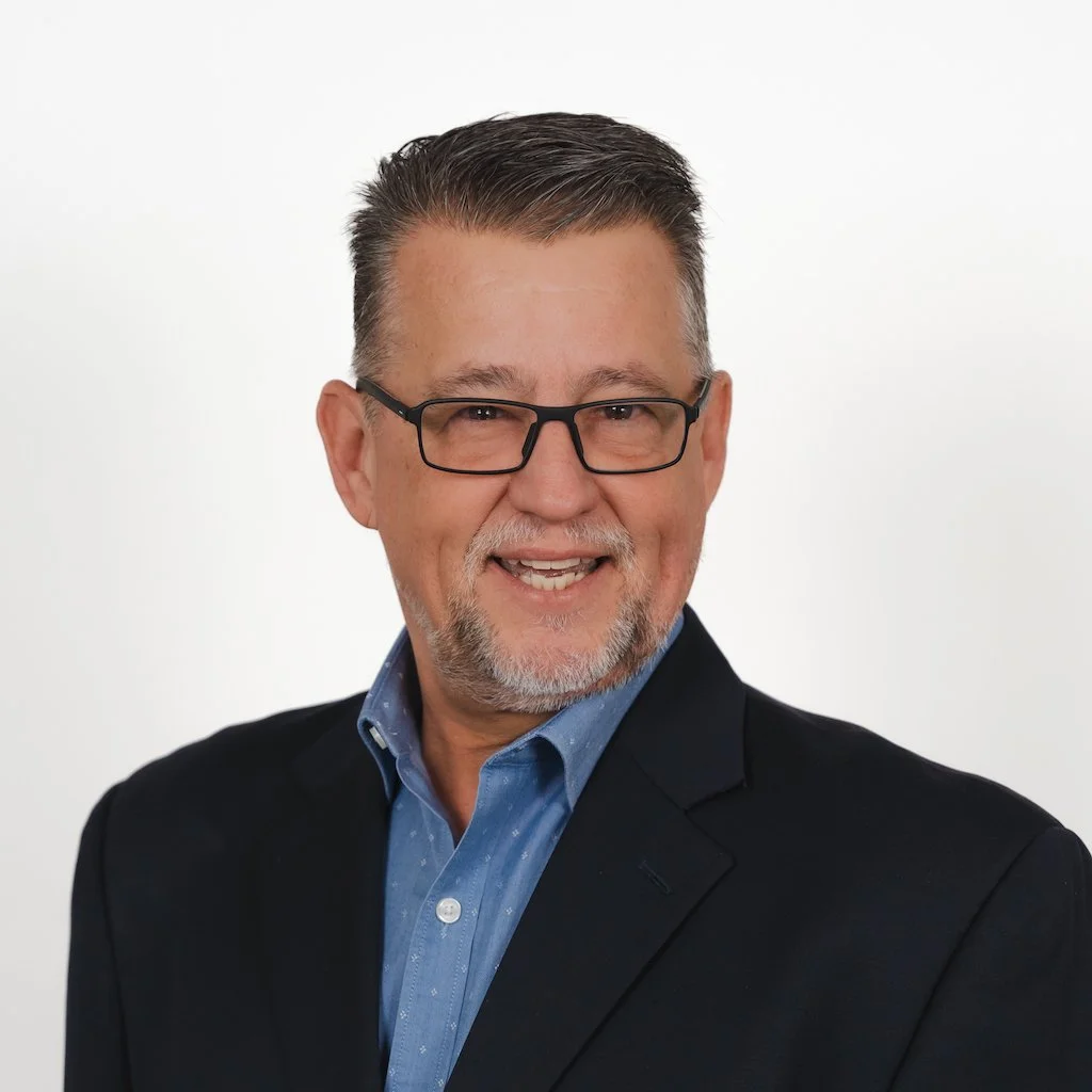 A middle-aged man with glasses, a short beard, and gray hair, smiling and wearing a black suit jacket over a blue dress shirt, against a plain white background.