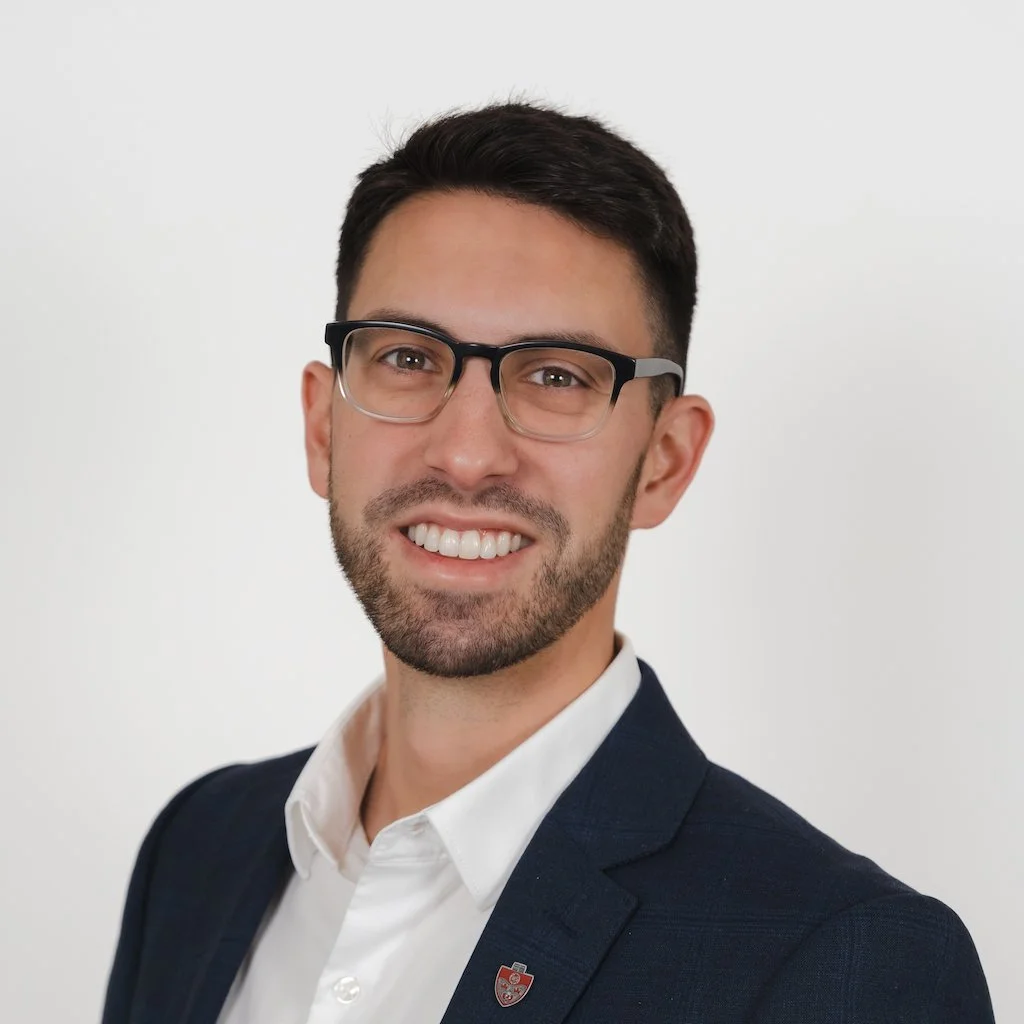 A man with dark hair, glasses, and a beard, smiling, dressed in a white shirt and a dark blazer, against a plain white background.