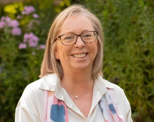 Smiling woman in glasses and white blouse outdoors with purple flowers and greenery in the background.