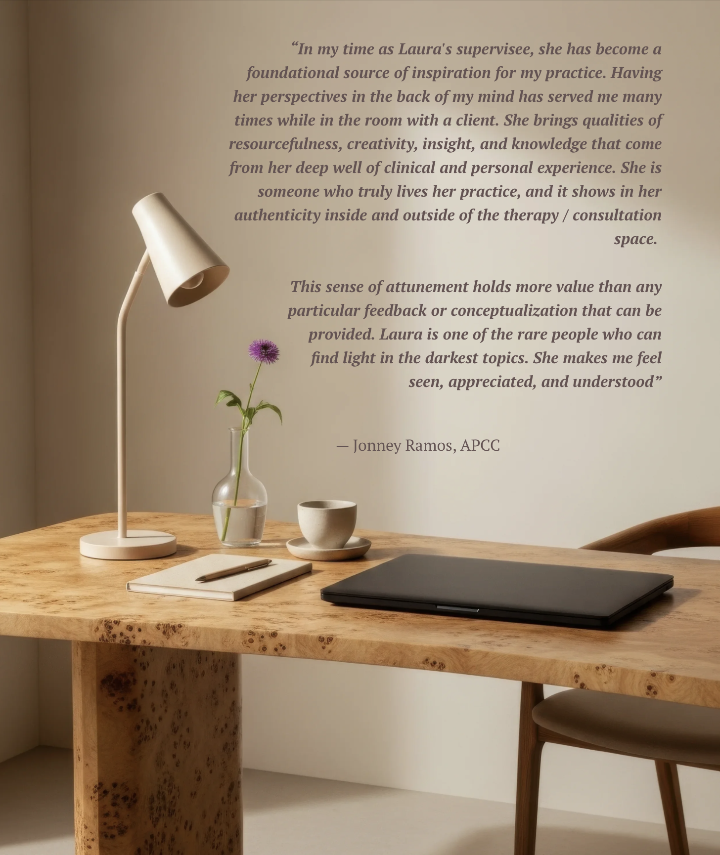 A wooden desk with a white adjustable desk lamp, a small glass vase with a purple flower, a white cup and saucer, a closed black laptop, a notebook with a pen, and a chair. A wall behind the desk has a quote about Laura and her qualities.