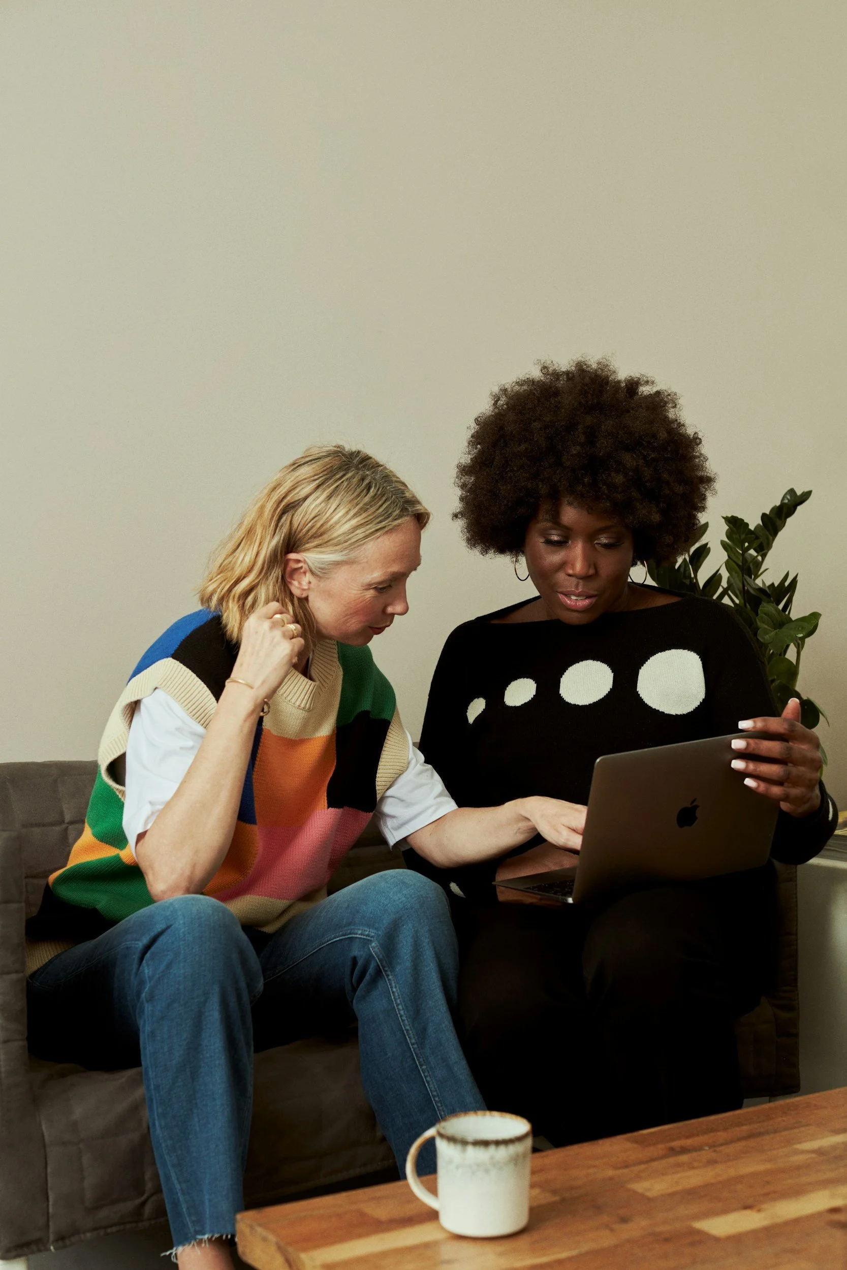 Two women sitting on a gray couch looking at a laptop screen together. One woman has curly hair and wears a black sweater with white circle patterns, the other has blonde hair and wears a colorful striped sweater. A coffee mug is on a wooden table in front of them. There is a plant in the background.