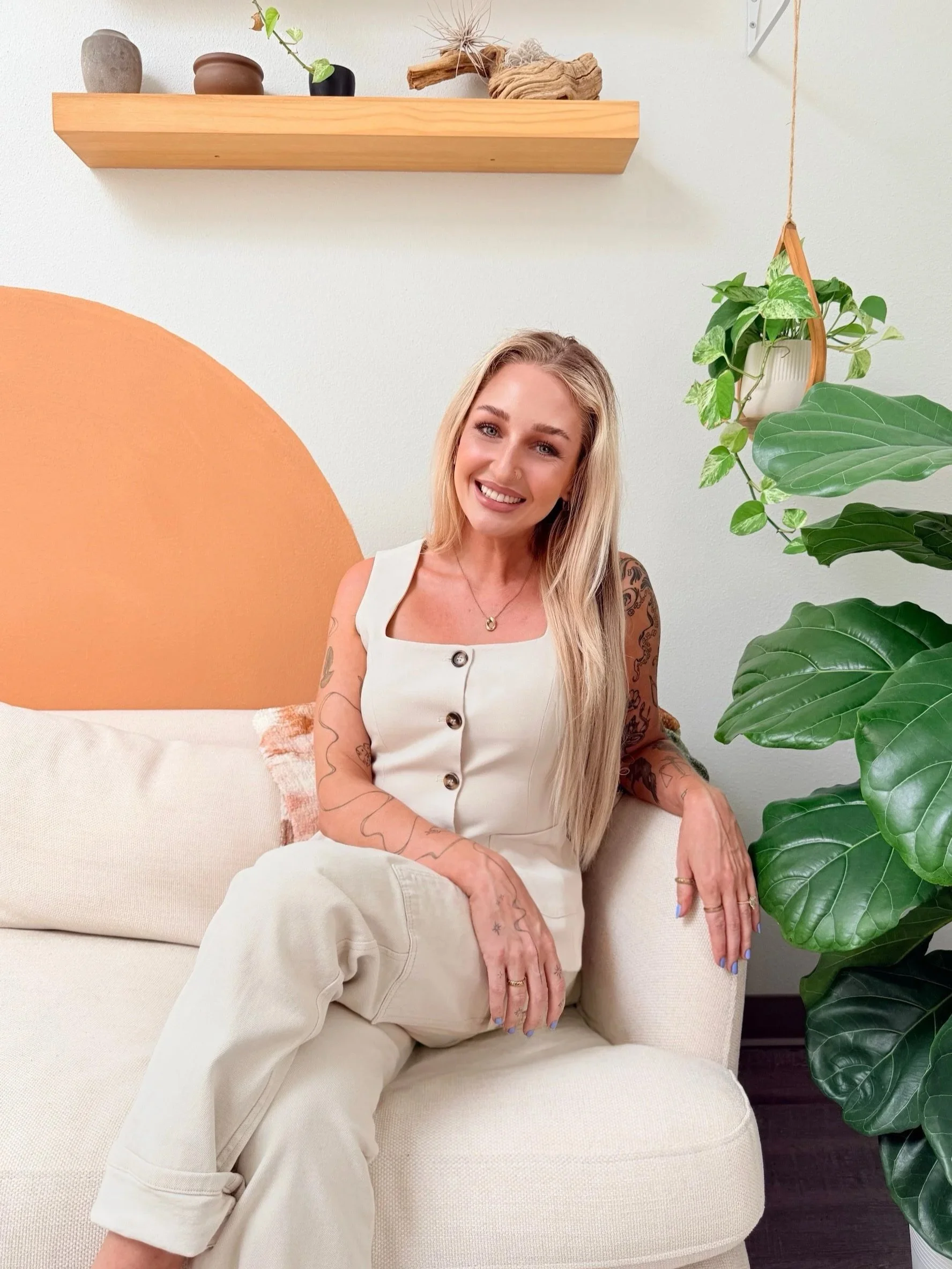 A smiling blonde woman with tattoos on her arms, wearing a white tank top with buttons, seated on a beige sofa with her legs crossed. She is in a room with a white wall, wooden shelf with decorative objects, and green plants, including a large leafy plant to her right.