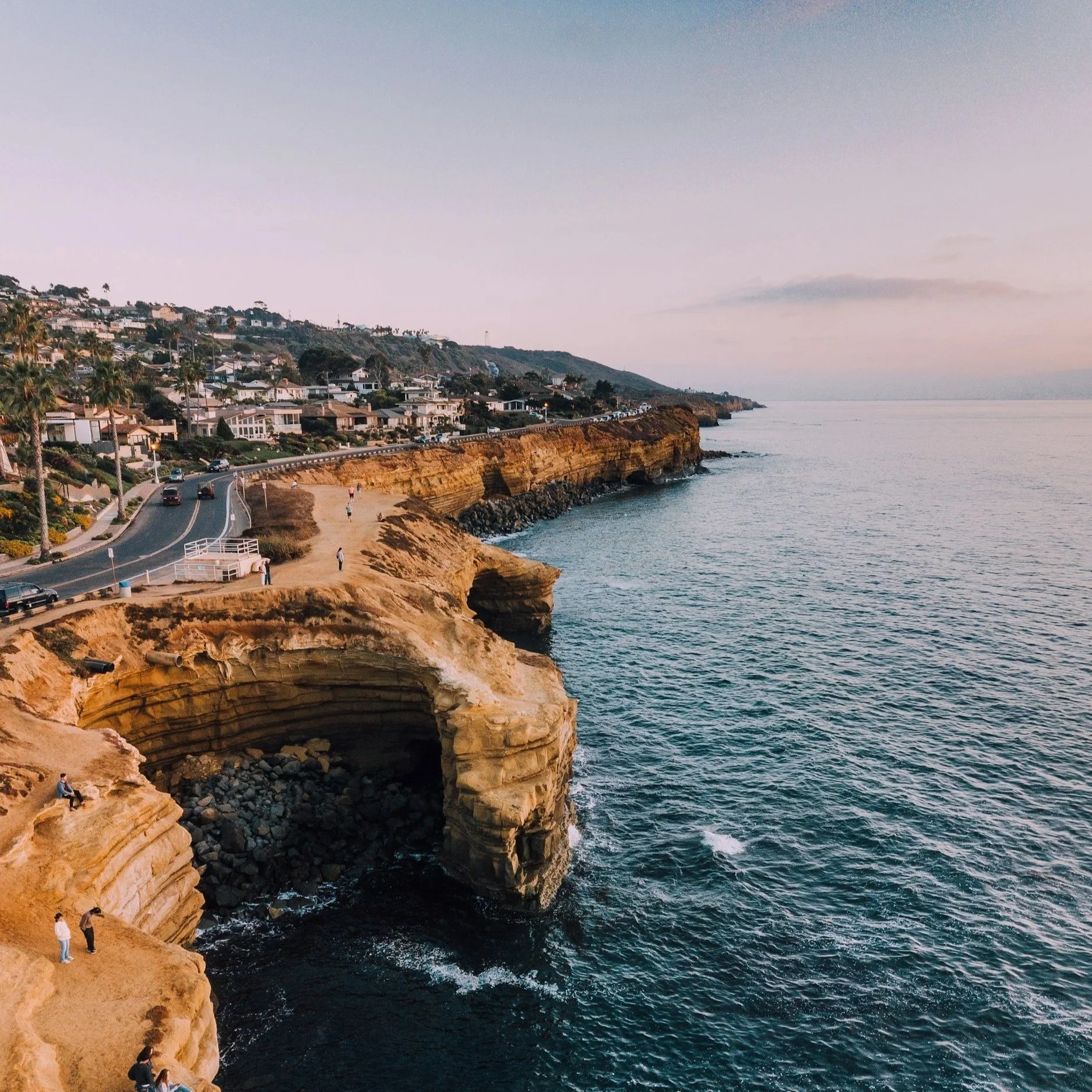 San Diego, California coastal cliffs with people walking and taking photos, residential houses along the hillside, ocean waves hitting the rocks, and a sunset sky.