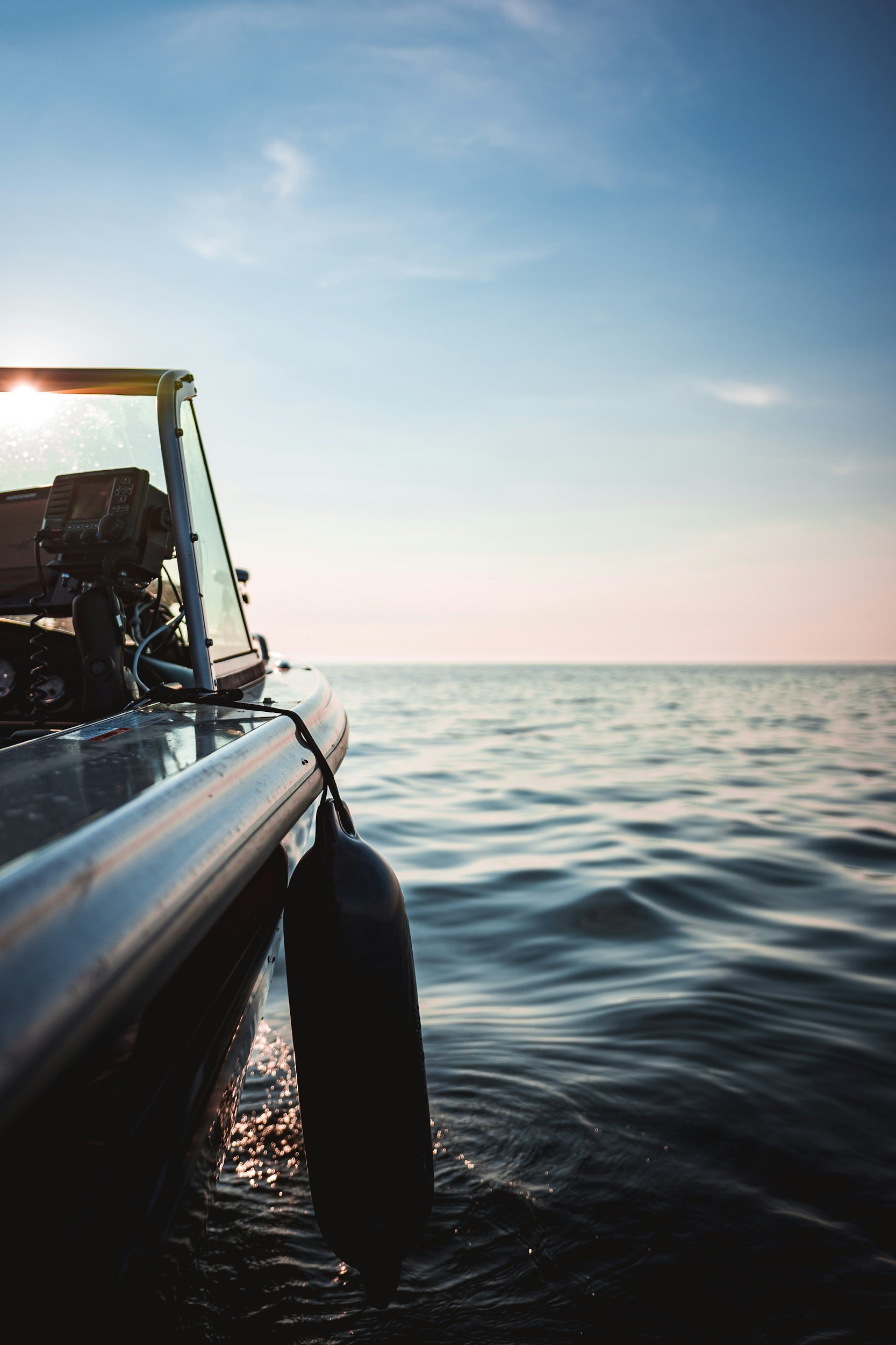 Close-up of a boat's edge with a fishing rod holder and a black fender, on a calm body of water with a clear sky at sunset or sunrise.
