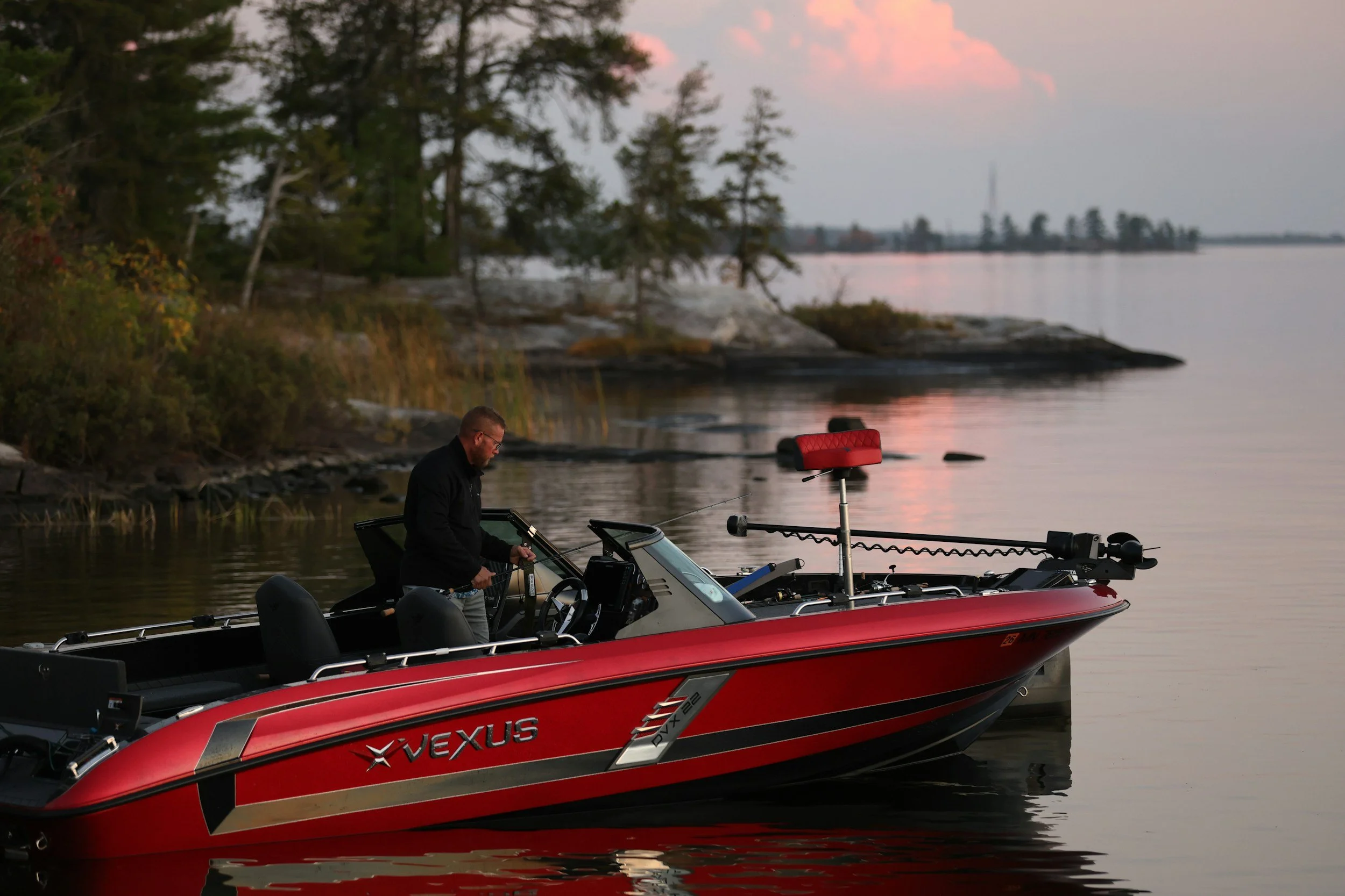 A man standing in a red Vexus bass boat preparing fishing equipment on a calm river during sunset, with trees and a distant shoreline in the background.