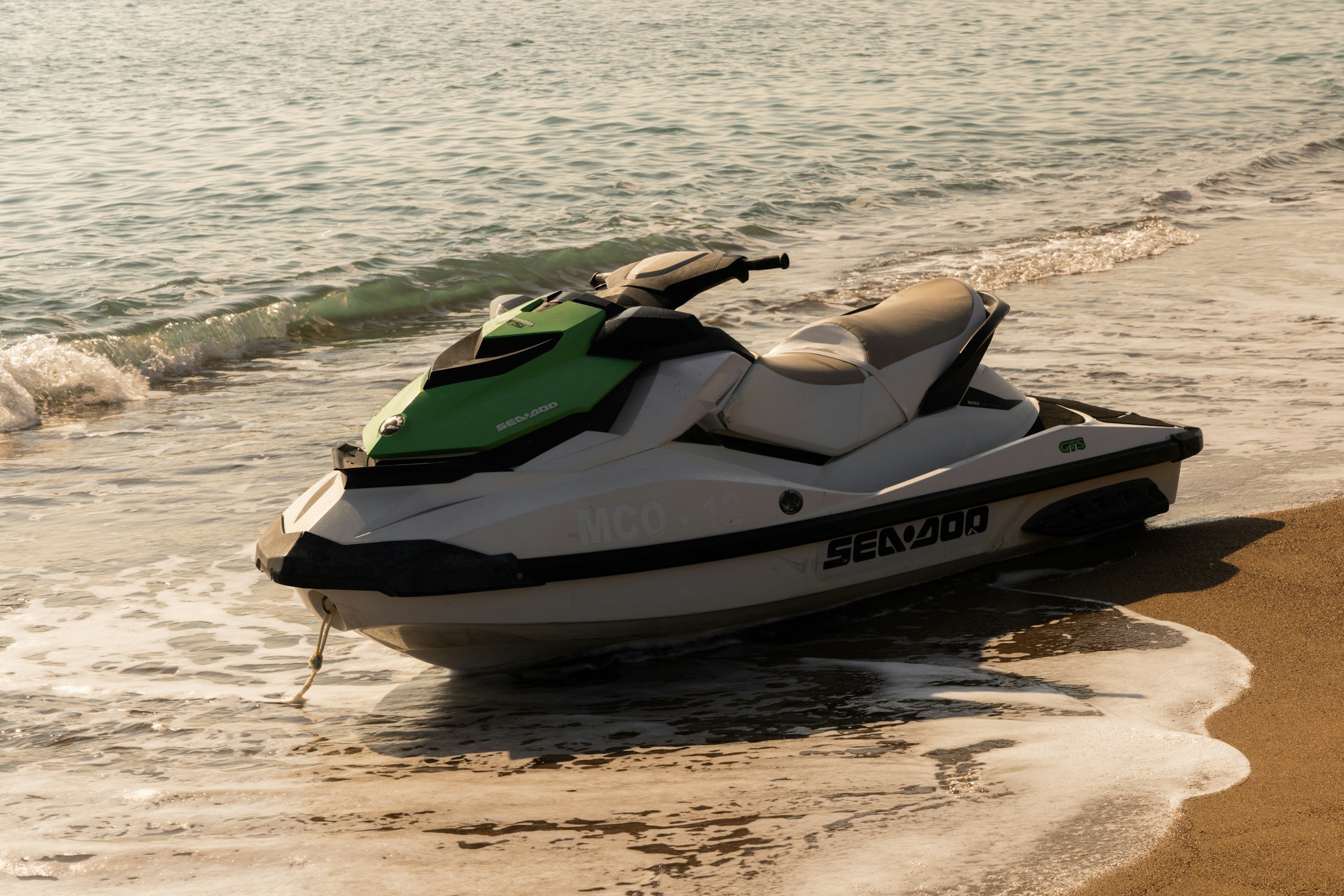 A green and white Sea-Doo jet ski resting on the sandy beach near the water's edge with small waves washing ashore.