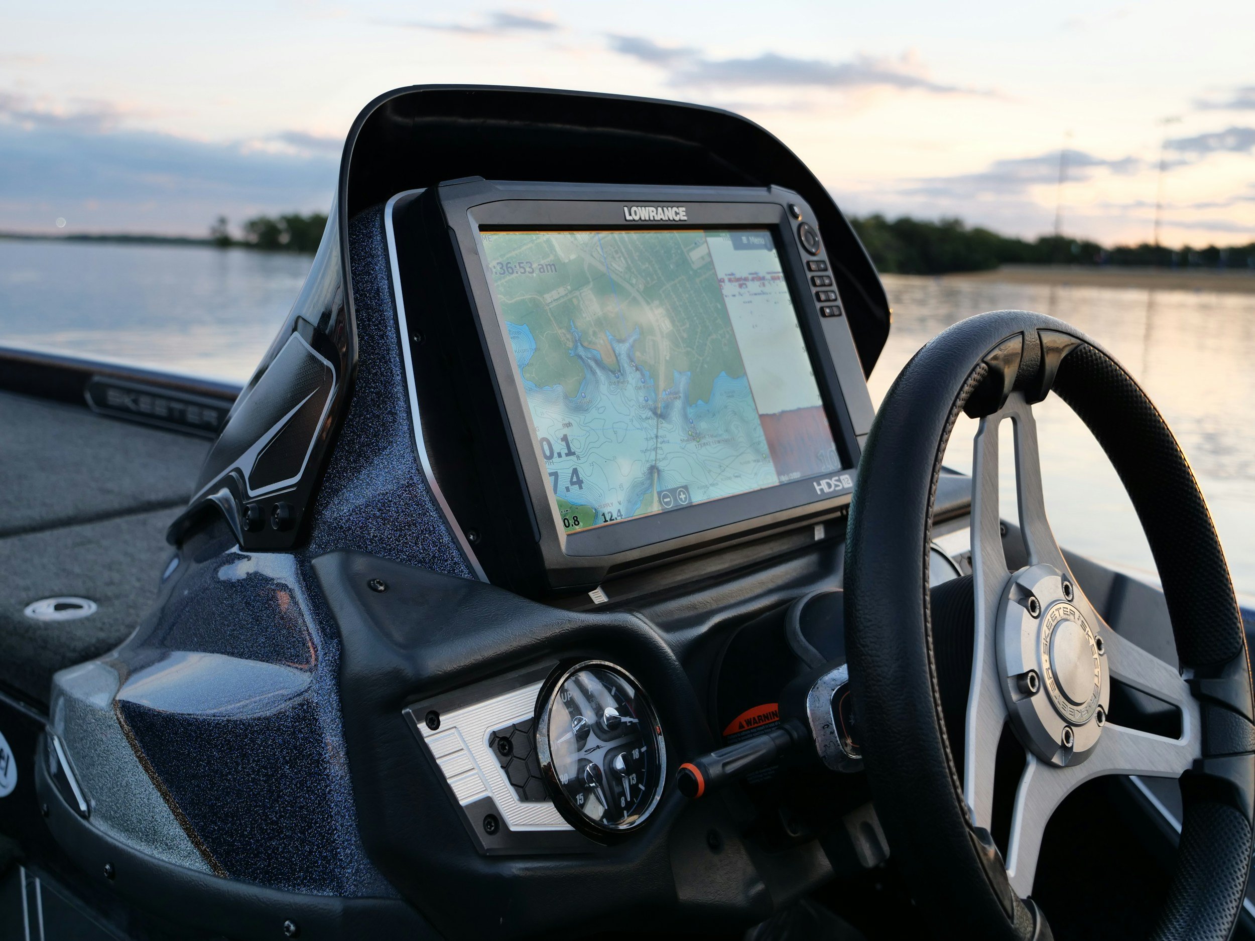 View of a boat dashboard with electronic navigation device and gauges, on a body of water at sunset.