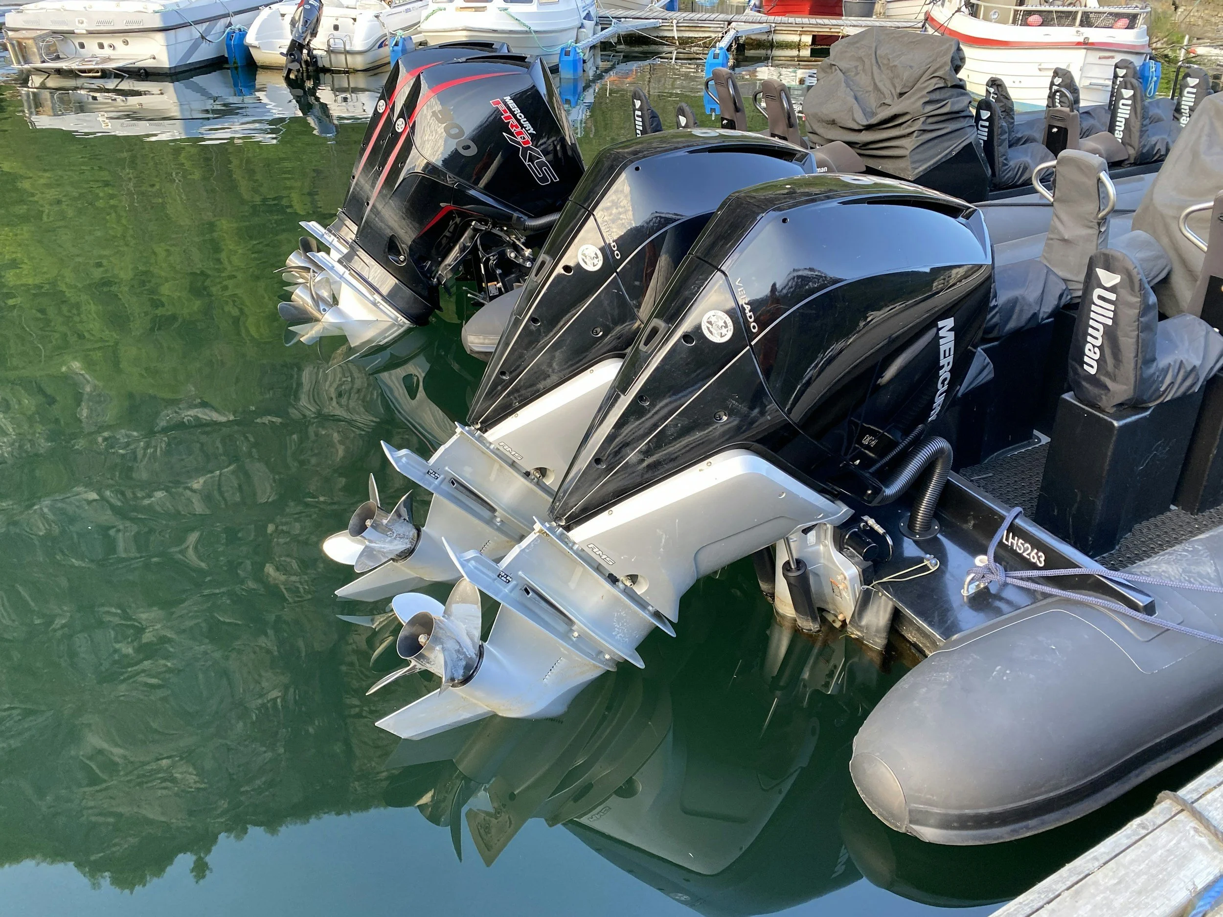 Two black Mercury outboard motors attached to a boat, with the reflection on the water and other boats docked nearby.