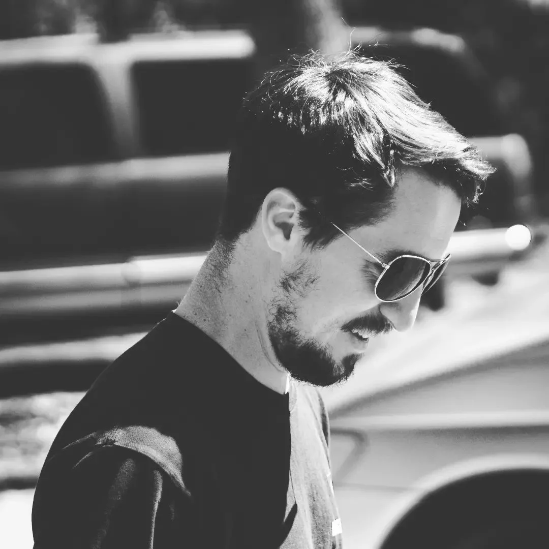 A black and white photo of a man with dark hair, wearing sunglasses and a beard, looking down and smiling.