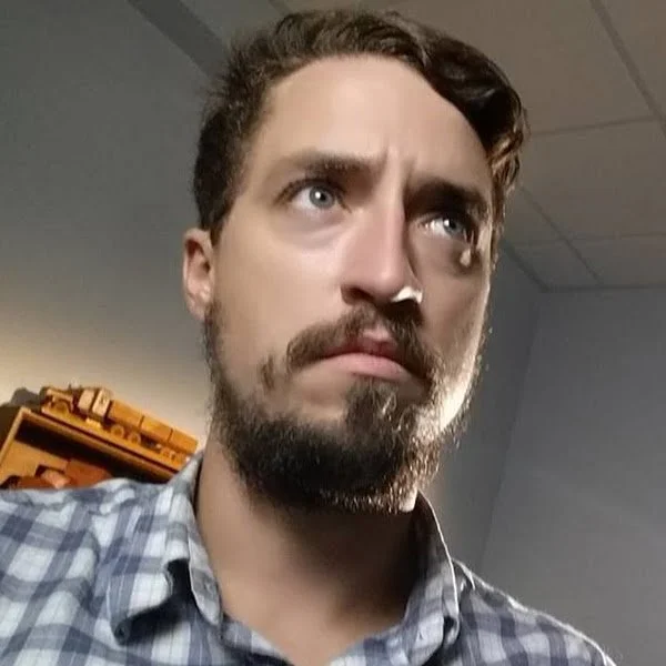 A man with brown hair, a beard, and mustache, looking upward with a serious expression, wearing a plaid shirt, in an indoor setting with a gray wall and a shelf in the background.