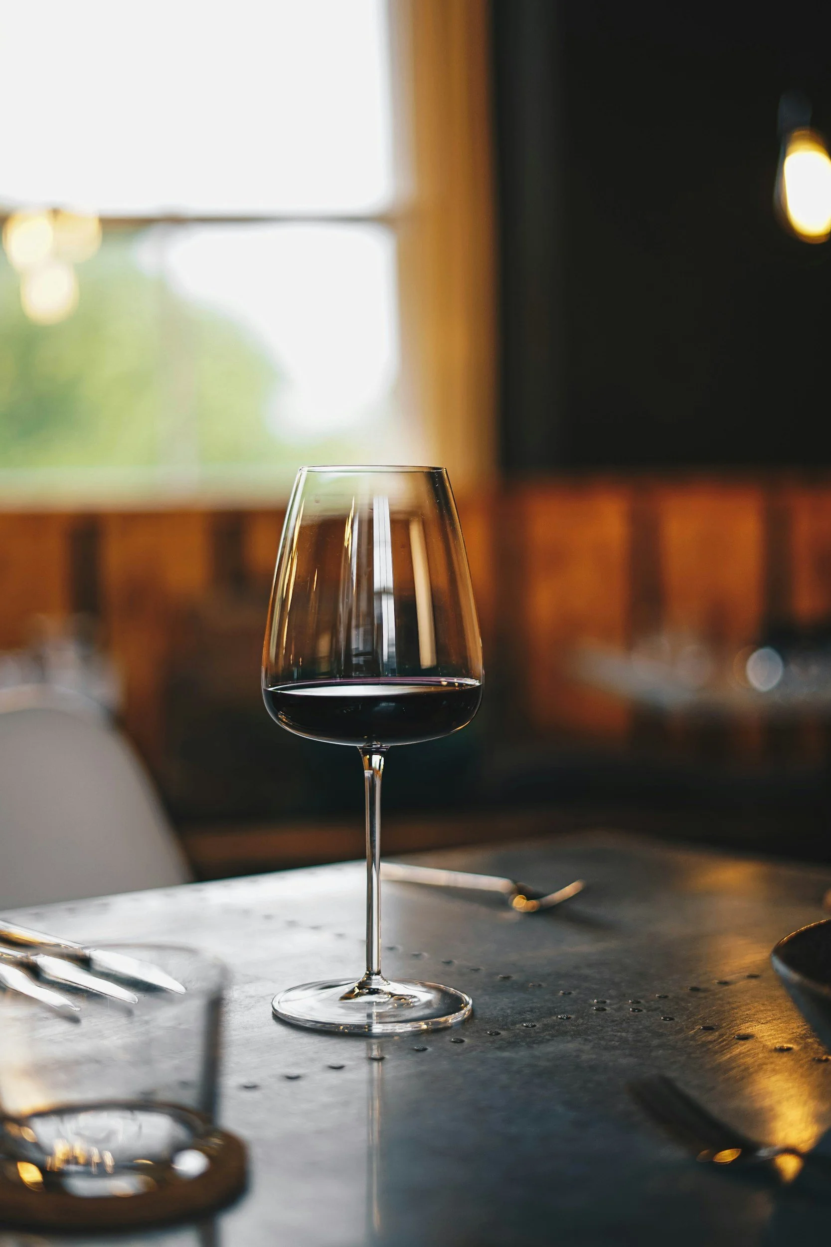A glass of red wine on a table with blurred utensils and a window in the background.