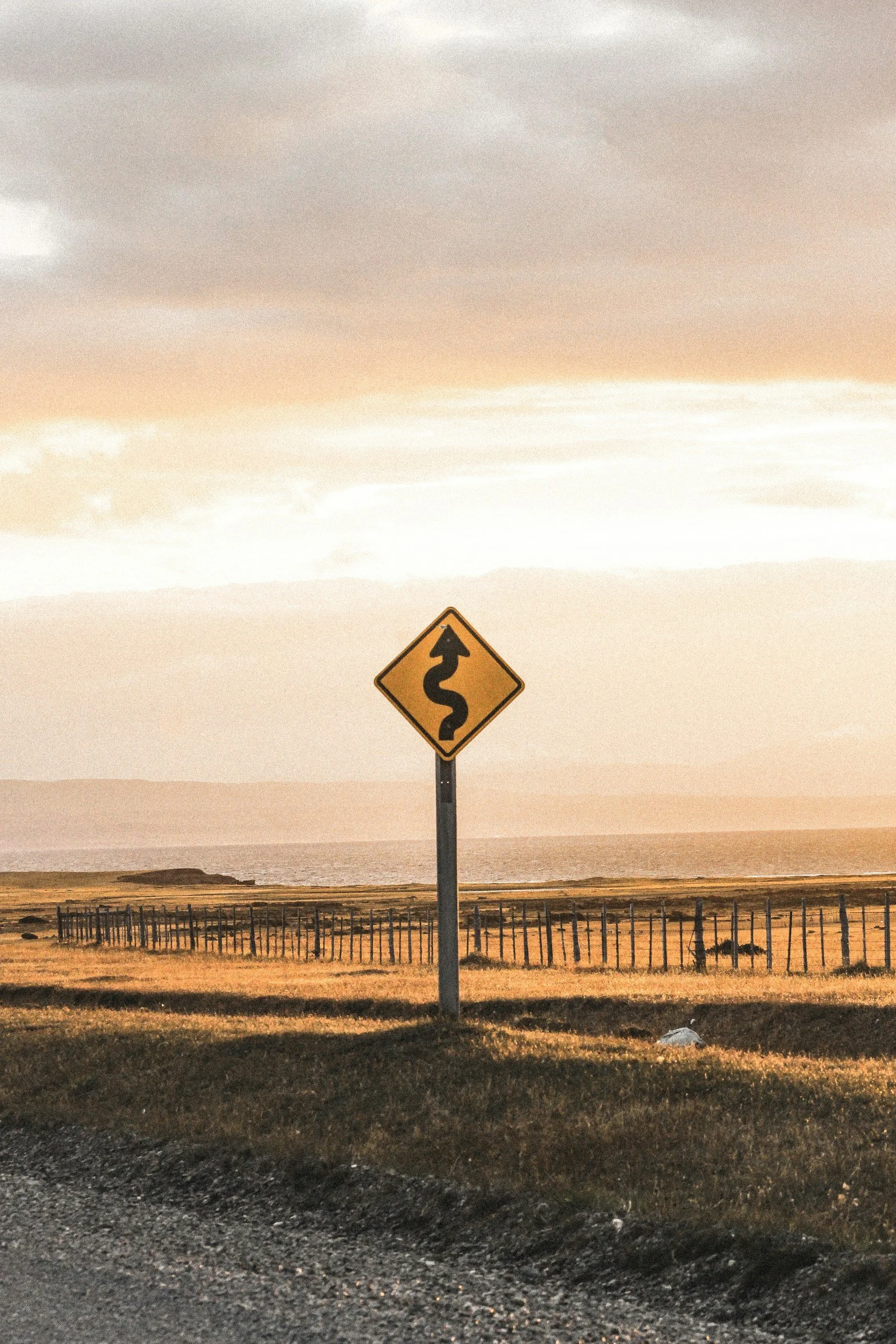 A warning road sign with a winding arrow is located near a coastline, with a wooden fence, fields, and an ocean under a cloudy sky at sunset.