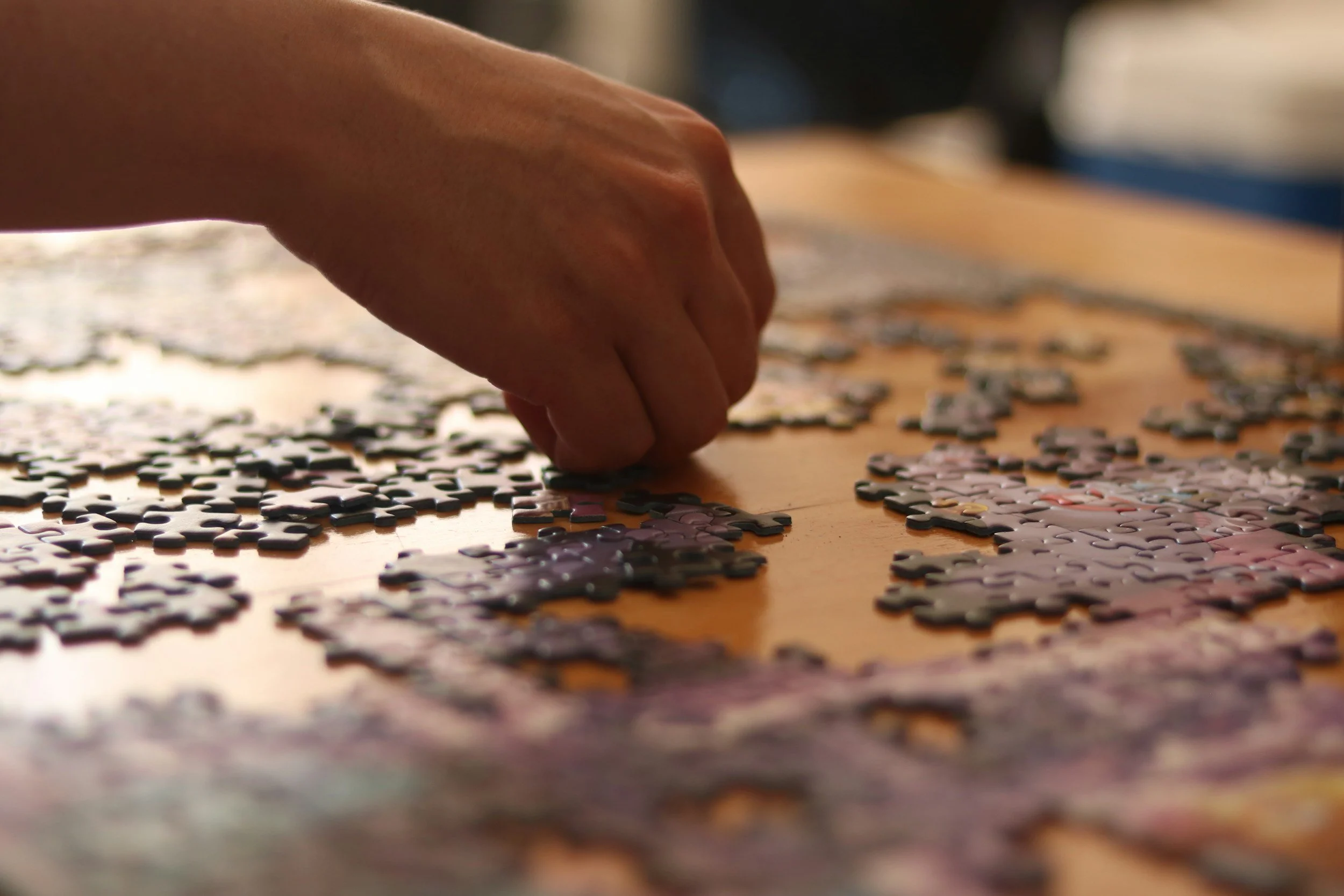 A person's hand working on a puzzle with small pieces spread out on a wooden surface.