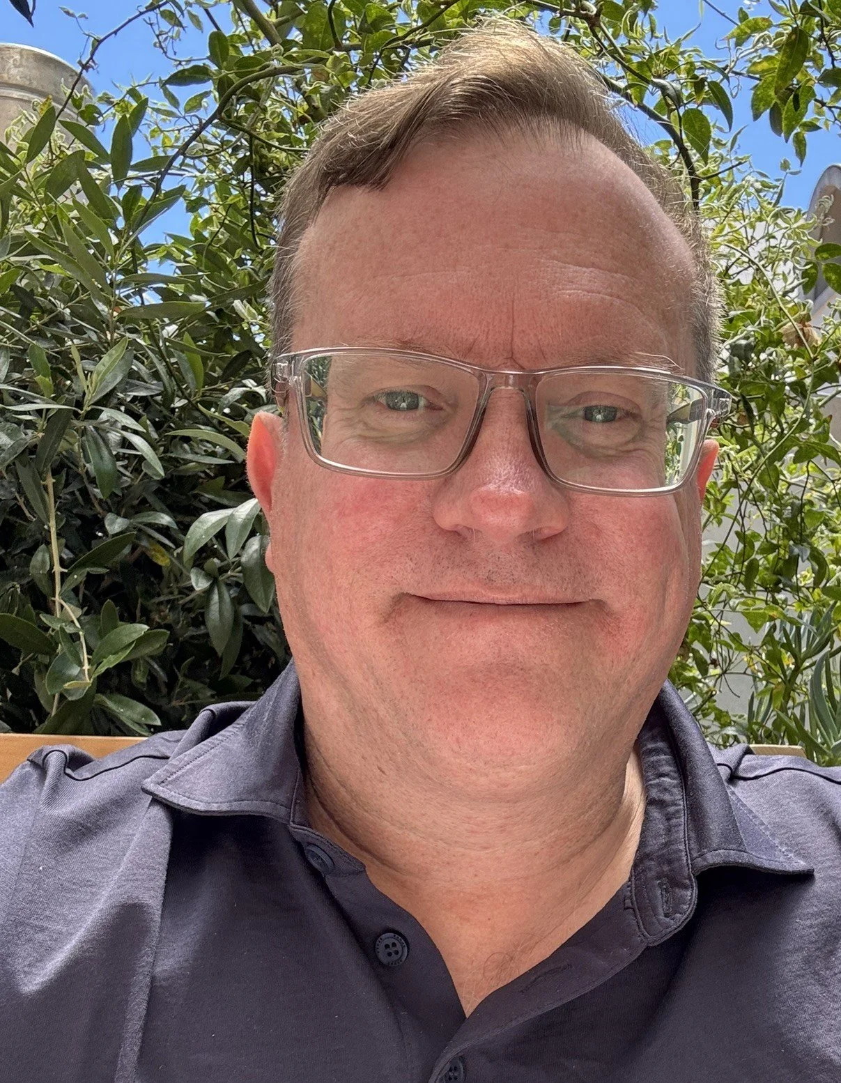 A man with short brown hair, wearing glasses and a dark collared shirt, smiling outdoors with green foliage and a clear blue sky in the background.