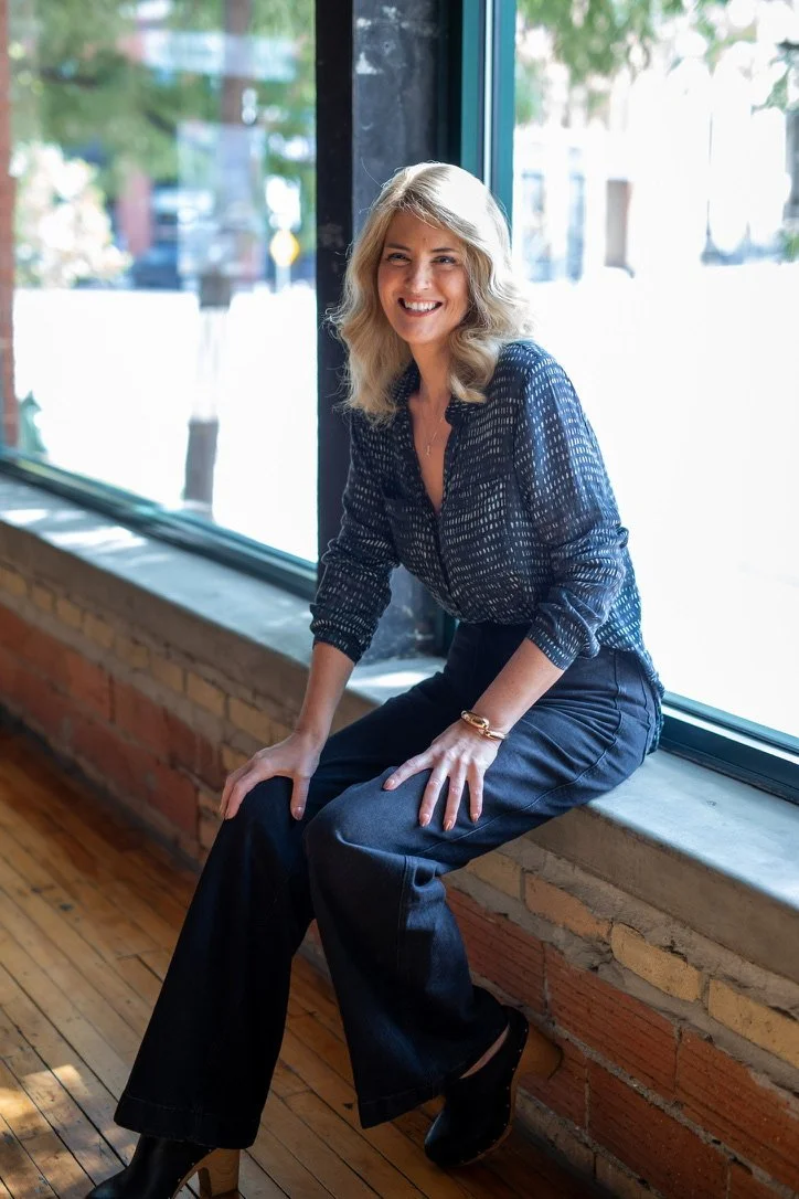 A woman with blonde hair, wearing a navy blouse with white dots, black wide-leg pants, and black heeled boots, sitting on a windowsill in a bright, modern space with wood flooring and brick walls, smiling at the camera.