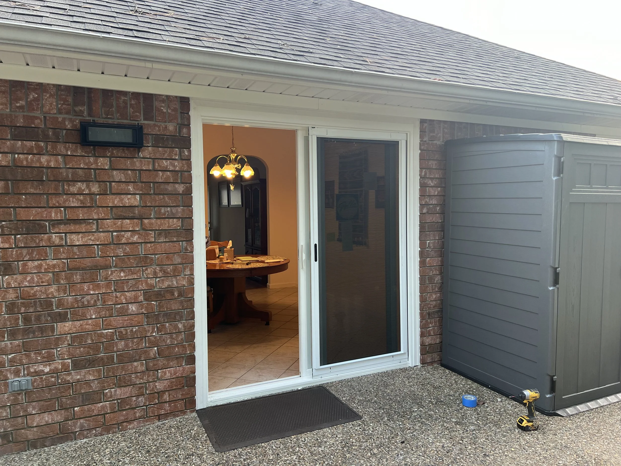 Outside view of a house with a brick exterior wall, glass sliding door, and a gray trash bin. The door opens into a room with a dining table and chandelier visible inside.