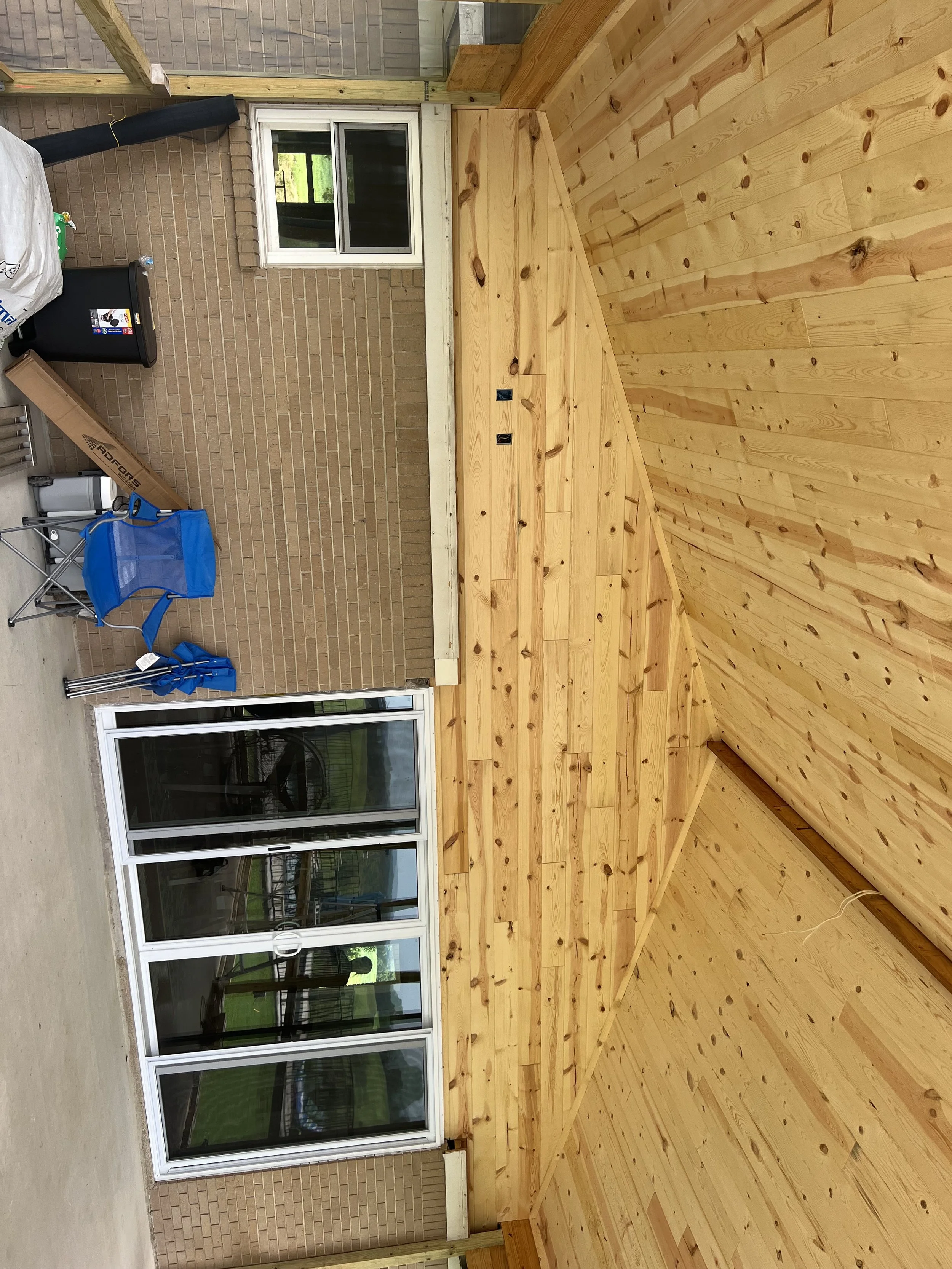 A partially finished wooden deck or porch area attached to a house. The deck is made of light-colored wood planks and has a sloped roof covering. There are sliding glass doors leading into the house, which has brick walls. Construction tools and mate