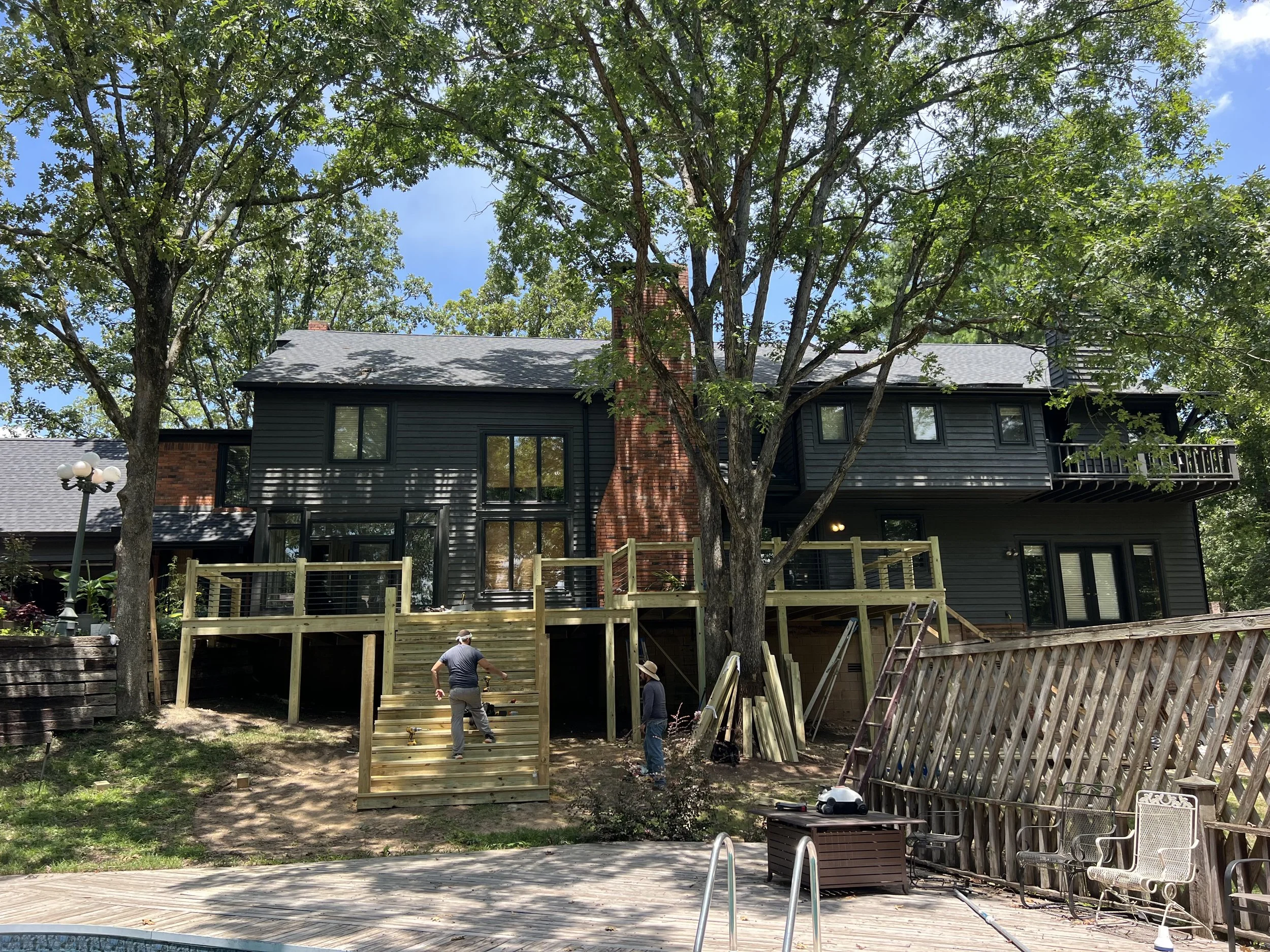 A house with black siding and a brick chimney is under construction in a backyard. There are several workers installing a wooden deck, and large trees with green leaves surround the area. A swimming pool with a metal ladder is visible in the foregrou