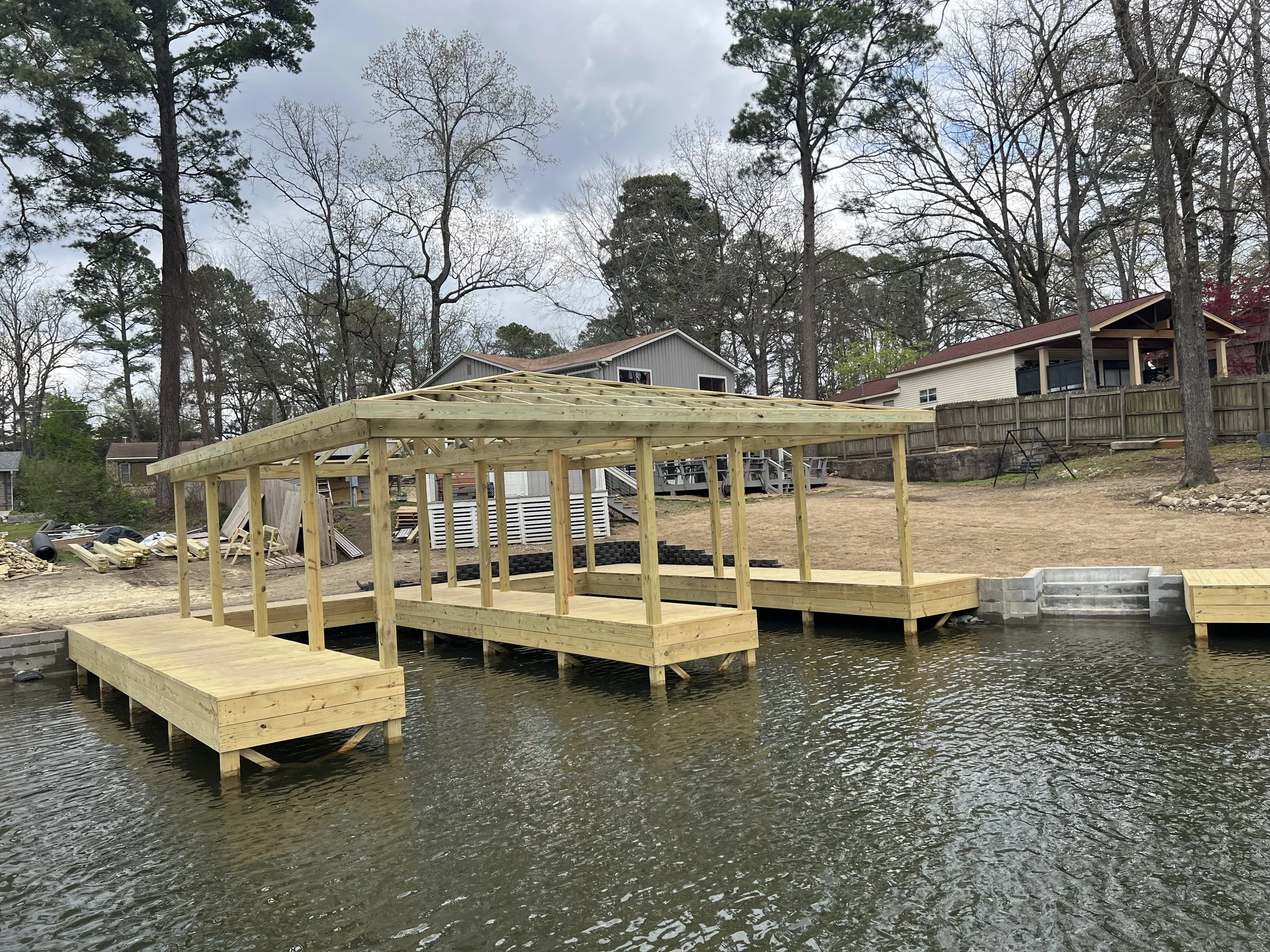 A wooden dock extending into a pond with an unfinished gazebo structure at the end, surrounded by a wooded backyard with houses in the background, under a cloudy sky.