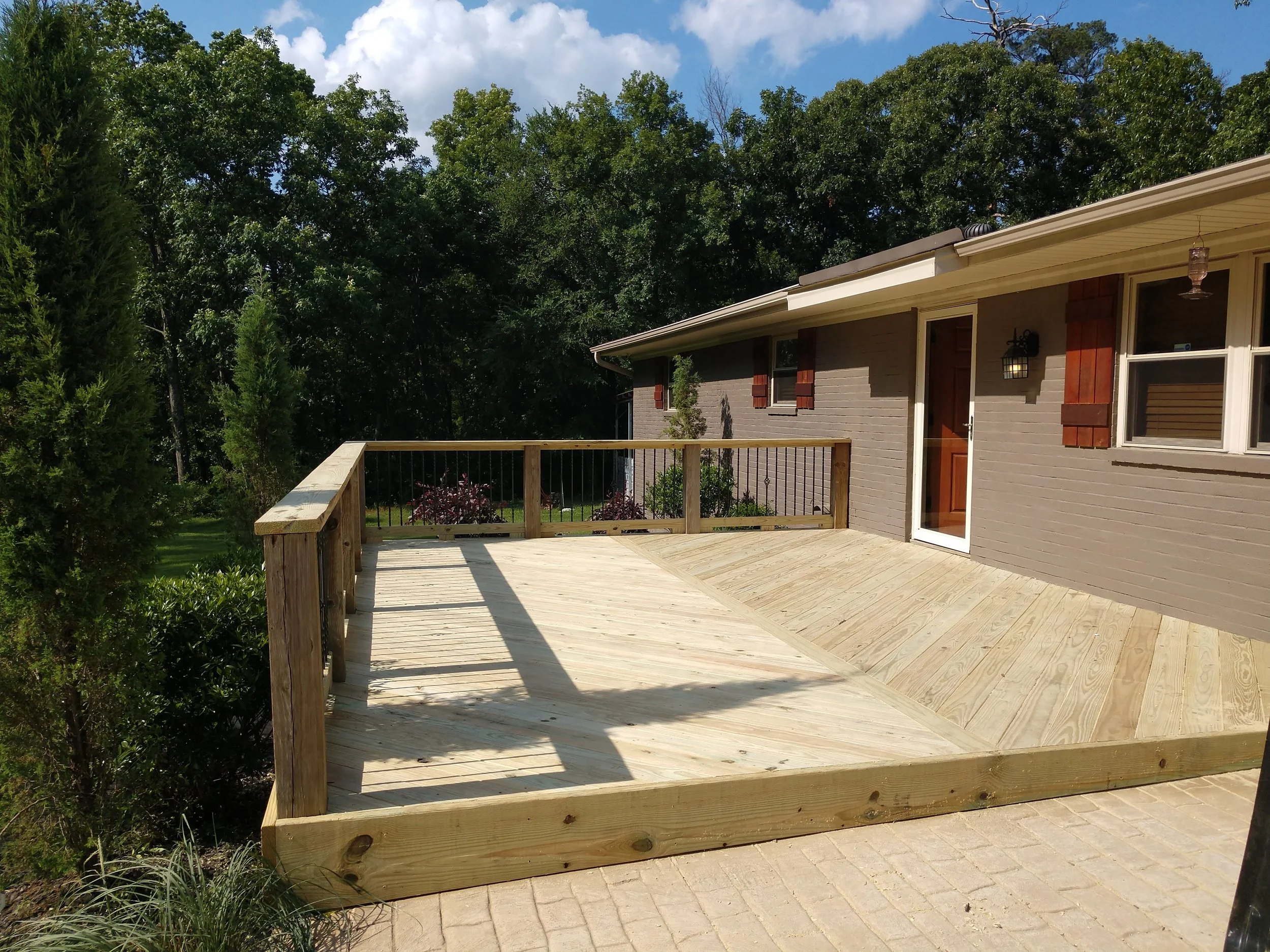 A newly built wooden deck attached to a house with a gray brick exterior, surrounded by green trees and bushes, under a partly cloudy blue sky.