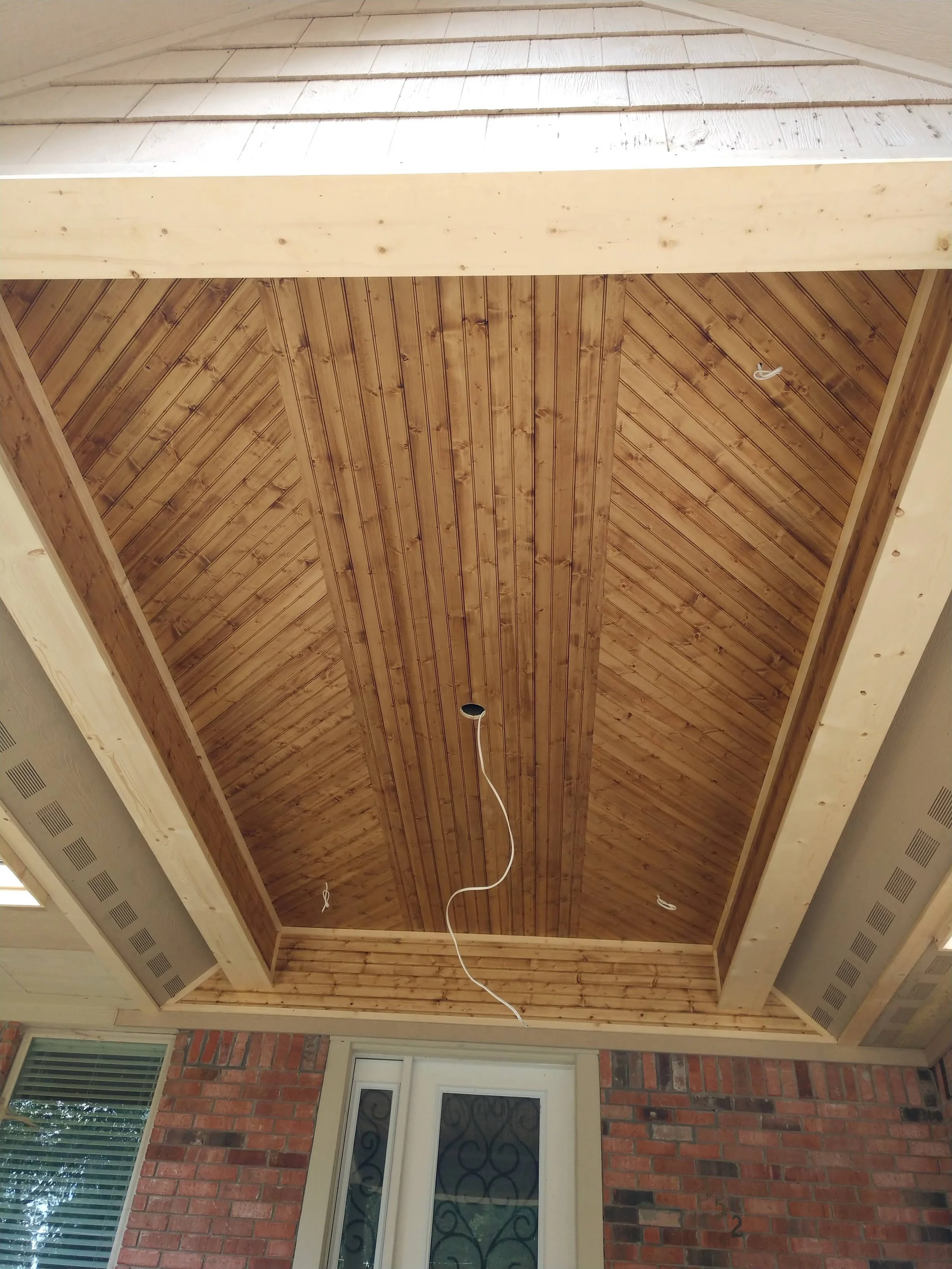 View of a wooden ceiling under construction in a house, with light-colored wood planks and visible white wires hanging down, and brick wall with a window below.
