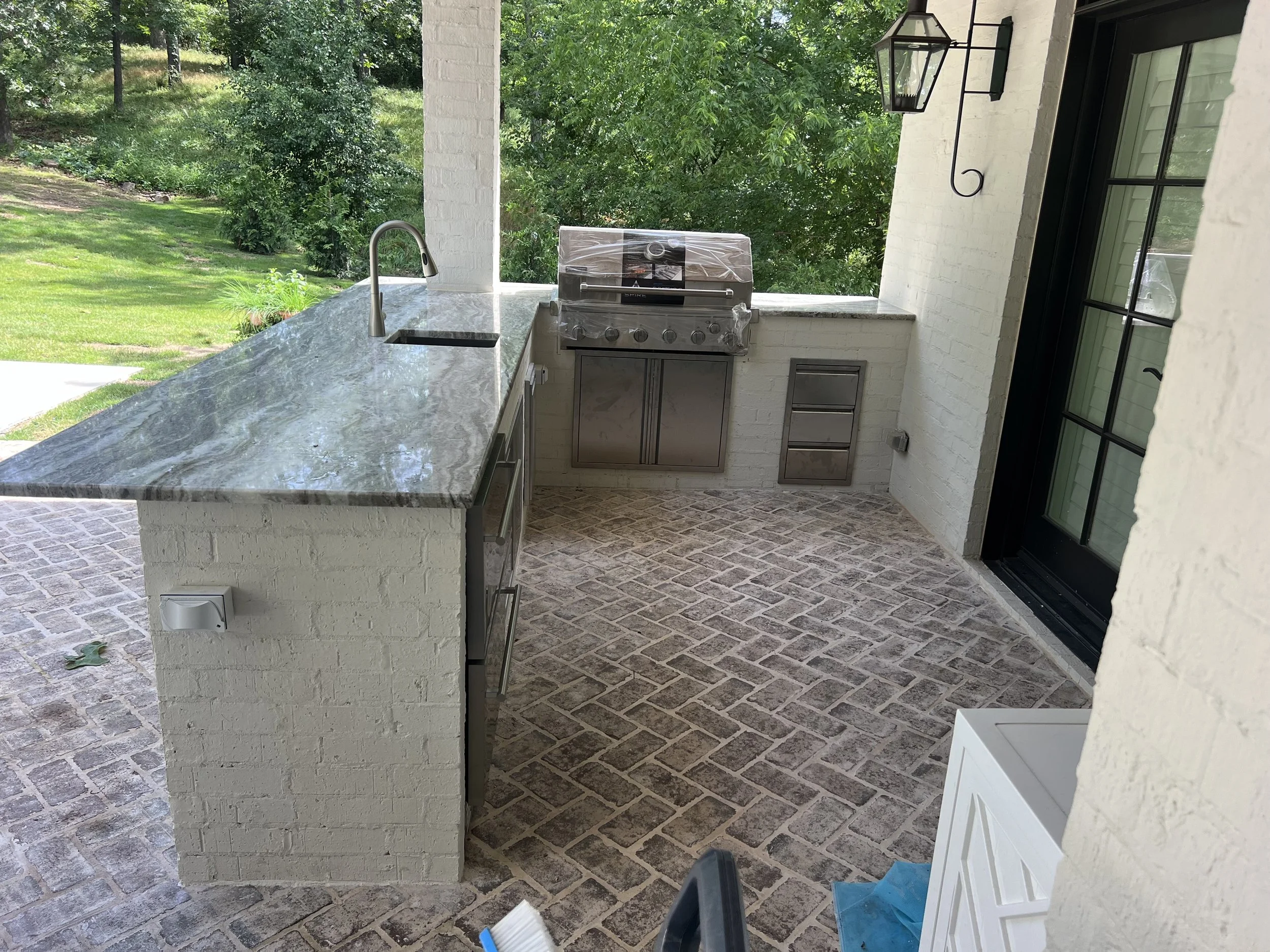 Outdoor kitchen area with a marble countertop, a grill, and a window. The area is surrounded by greenery and trees.