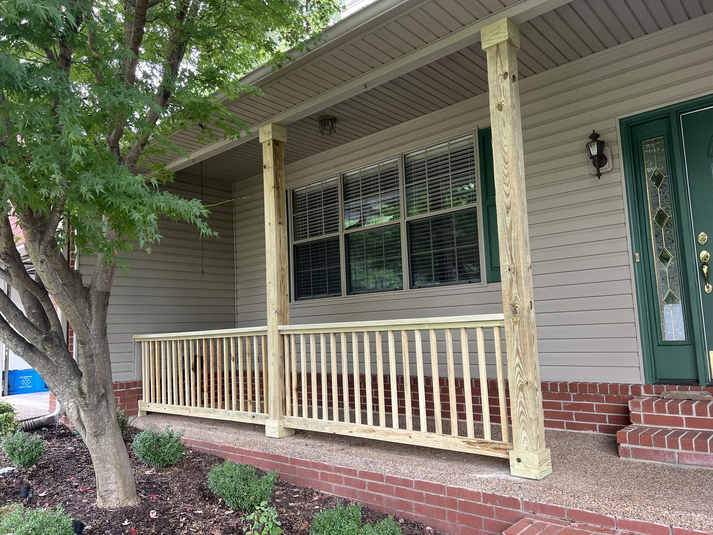 The front porch of a house with green shutters, a new wooden deck railing, brick steps, and a gray roof, with a large leafy tree in the foreground.