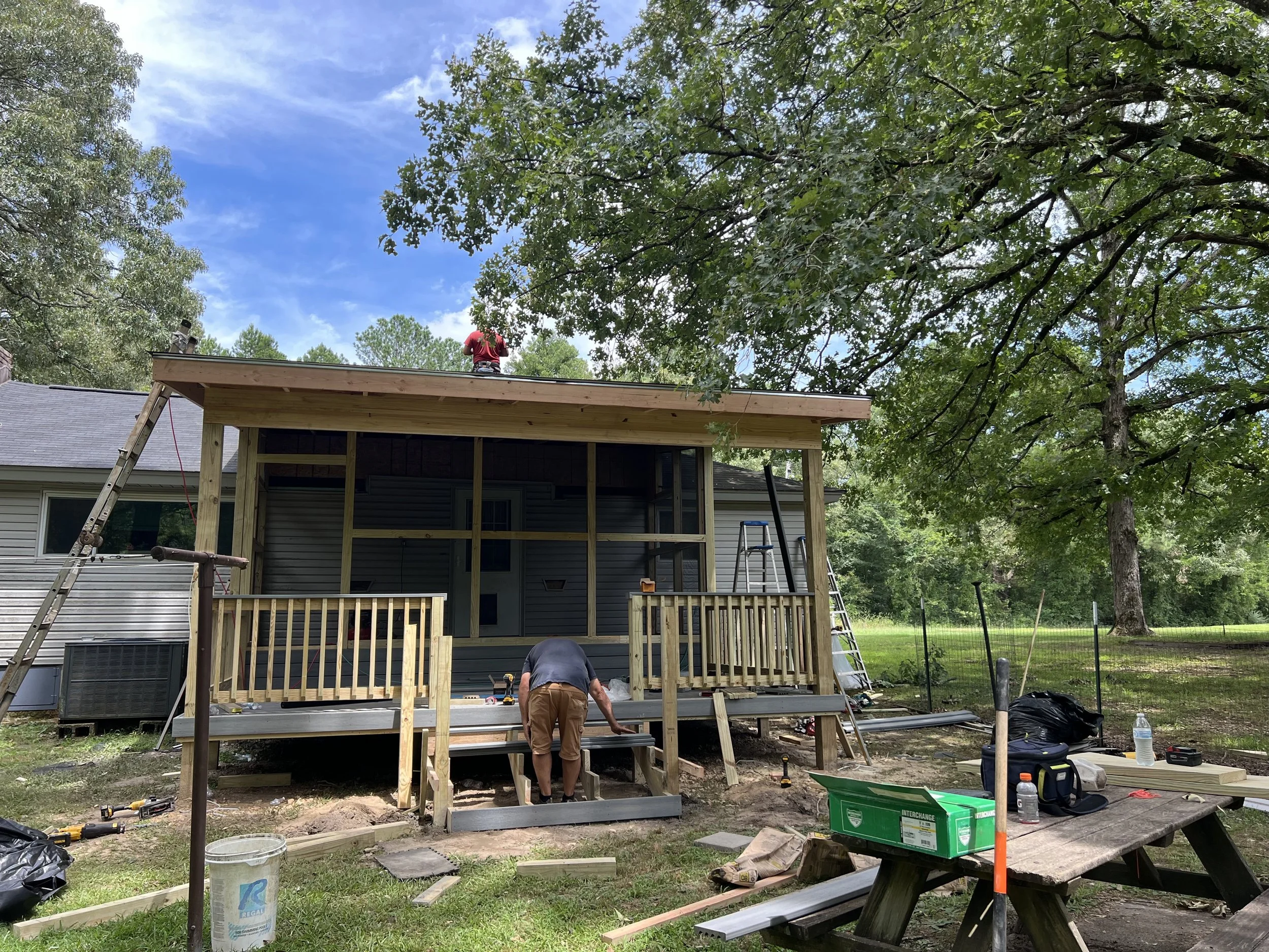 Workers building a deck extension on a house amid green trees and blue sky, with construction tools and materials scattered around.