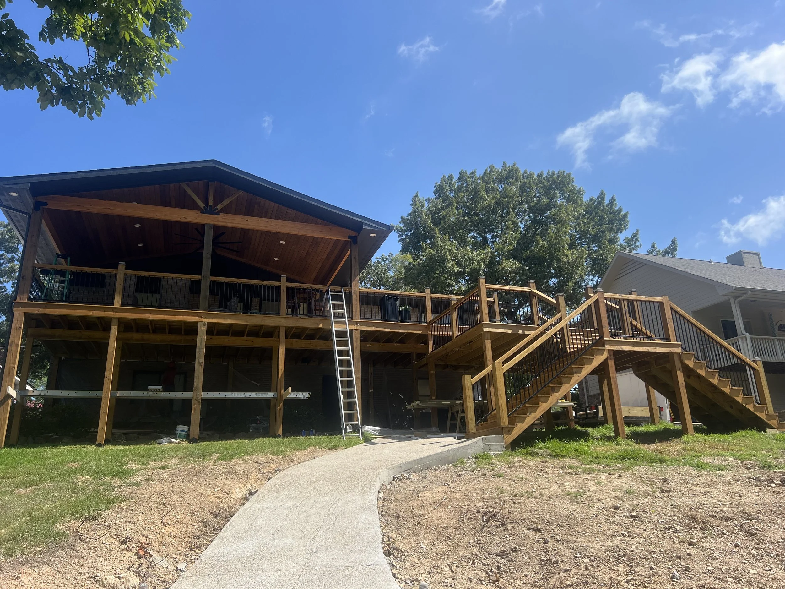Photo of a backyard deck under construction with a wooden staircase leading up to the deck. There is a ladder and construction materials underneath. The deck is partially built with a wooden railing and a covered roof area. The sky is clear with a fe