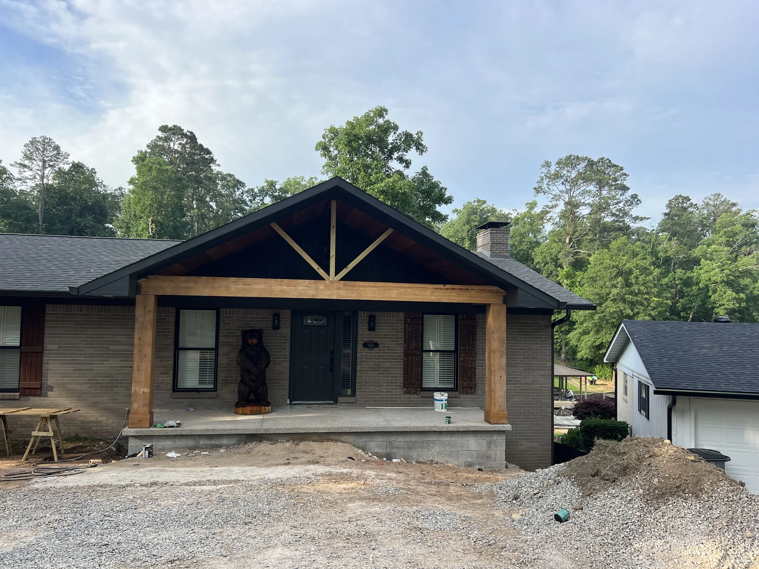 House under construction with a wooden porch frame, bear statue at the front, and a yard with construction materials and gravel, surrounded by green trees and a partly cloudy sky.