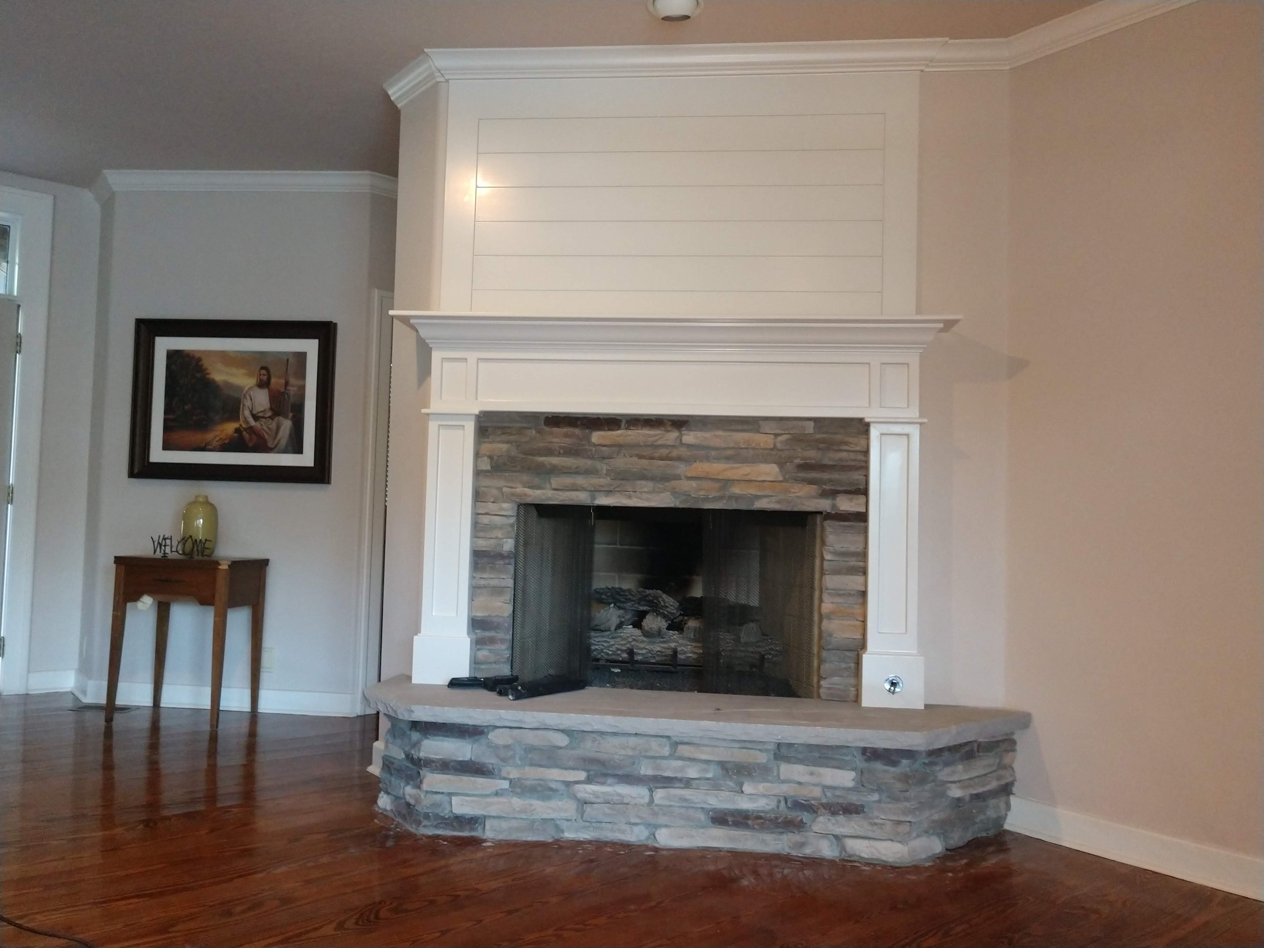 Living room with a stone and white fireplace, a wooden side table with a yellow vase and a framed picture of Jesus Christ, and hardwood floors.
