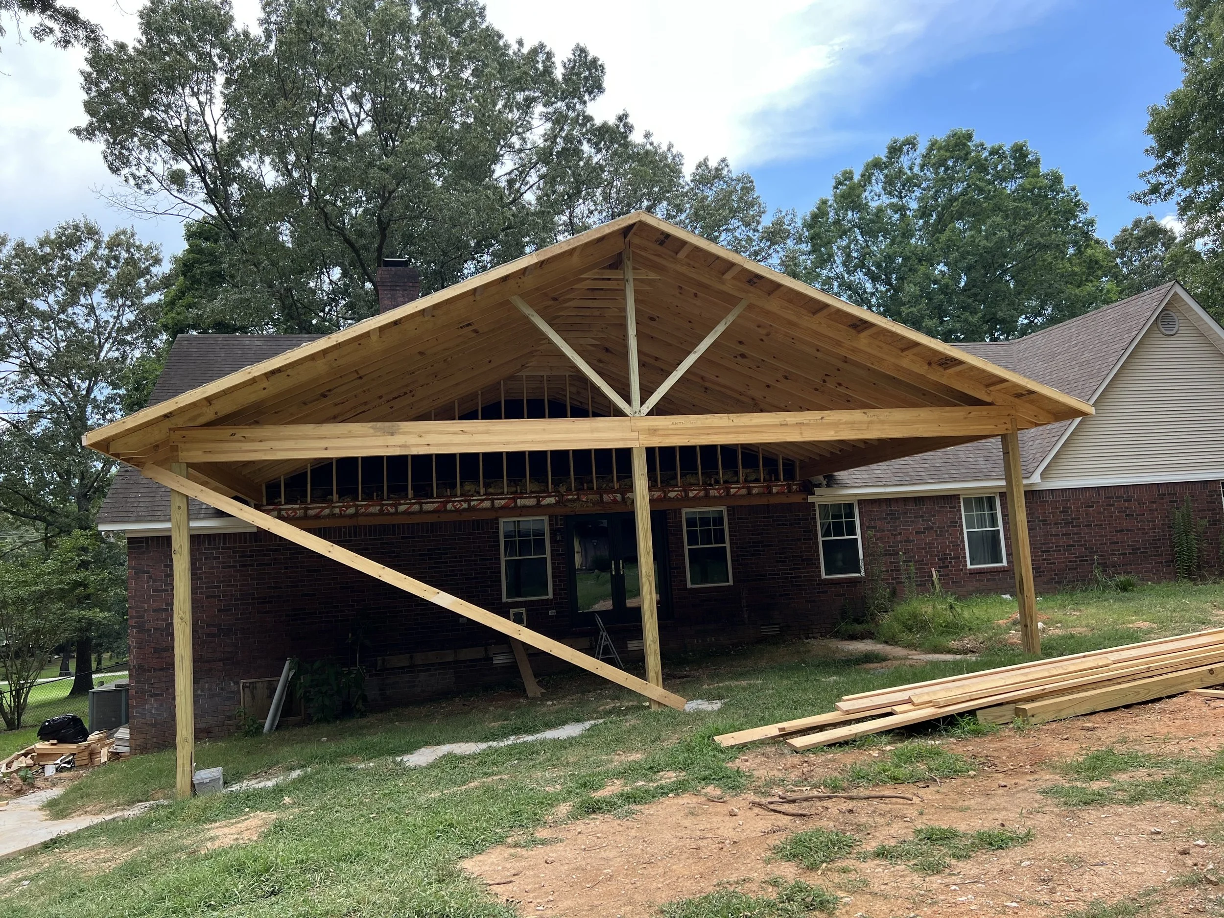A house under construction with a new wooden roof extension being built in the backyard, surrounded by trees and a blue sky.