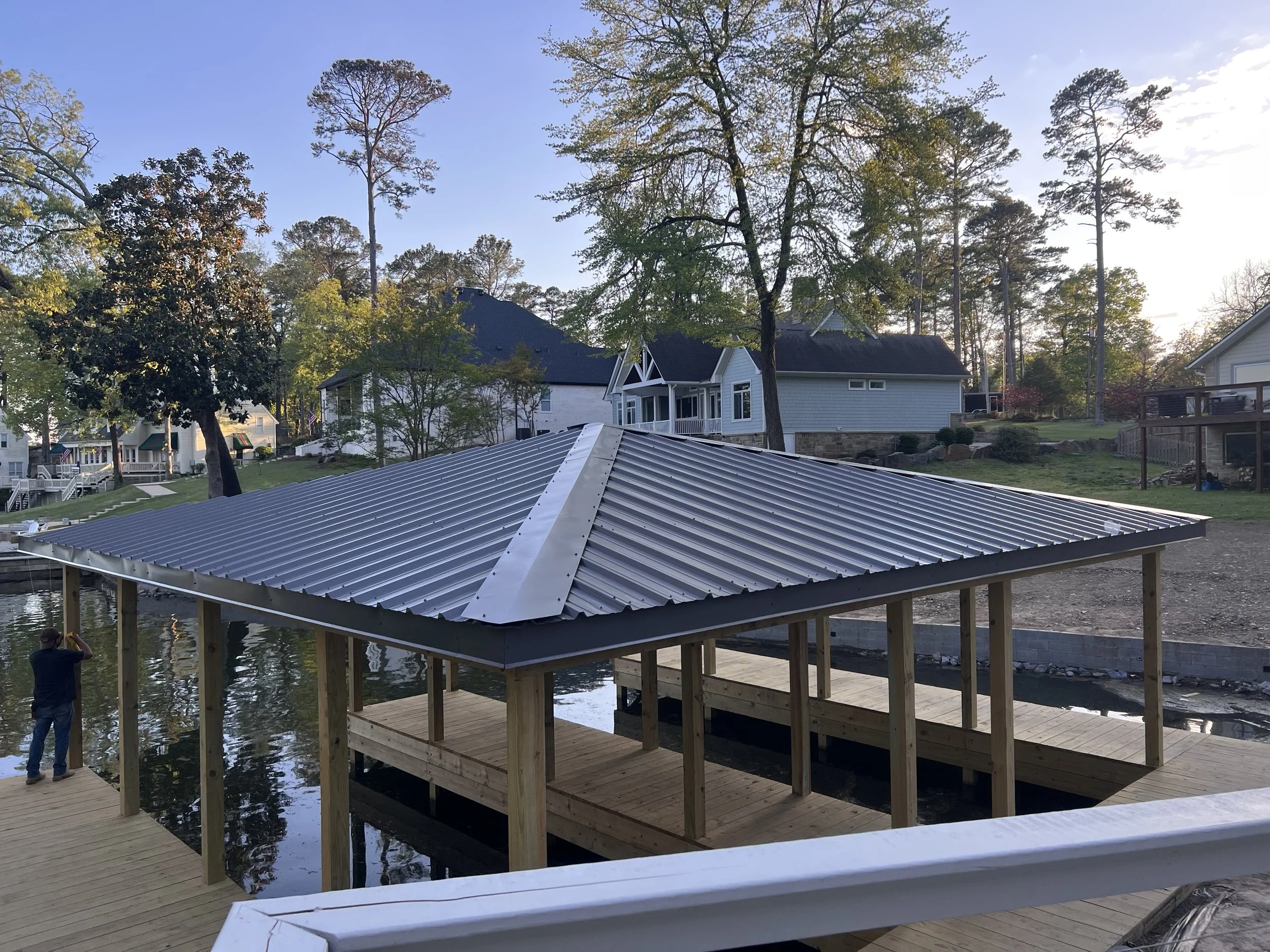 A wooden dock extending over a body of water with a house in the background, trees with autumn foliage, and a metal roofed structure in the foreground during late afternoon or early evening.