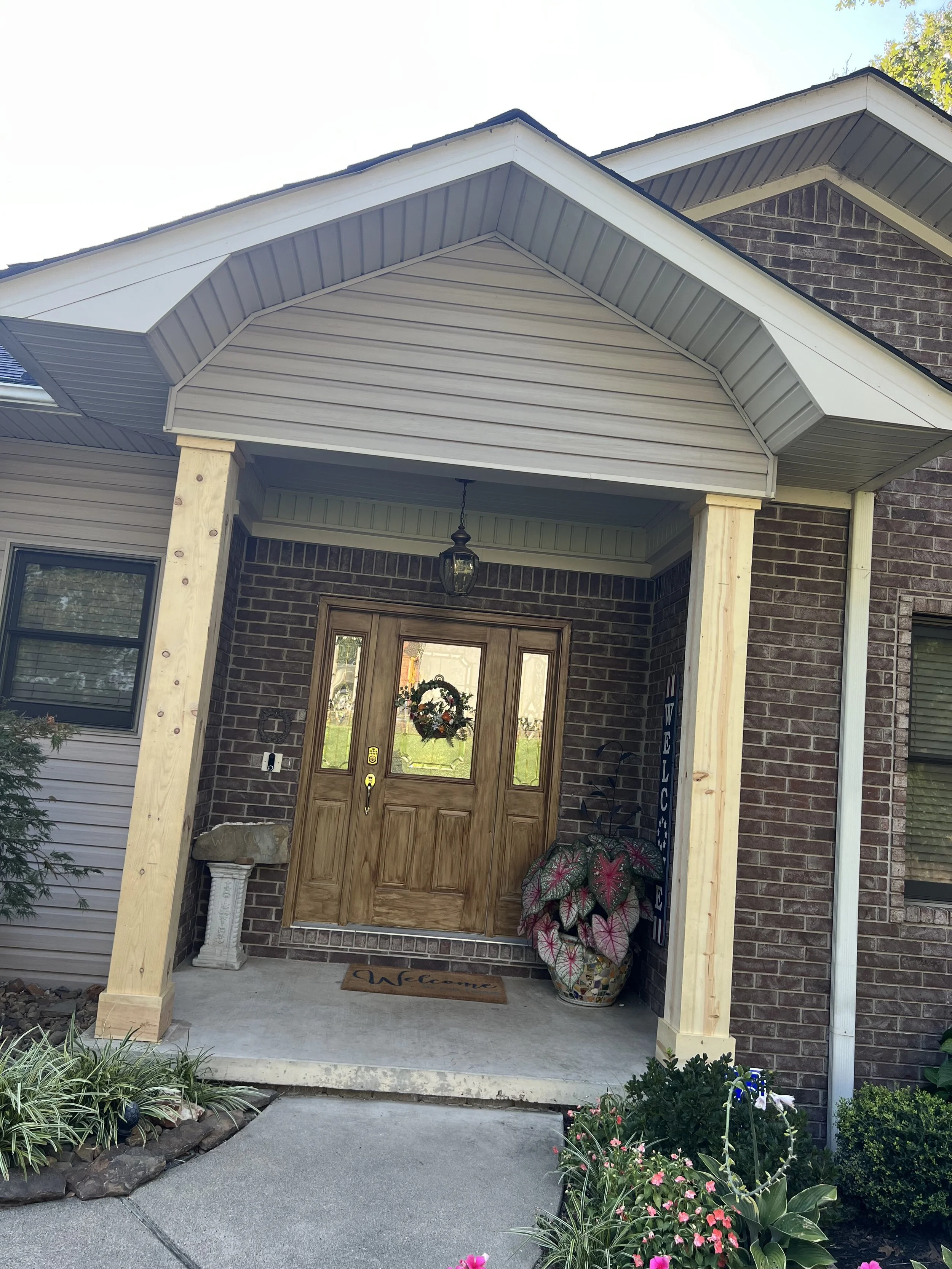 Front porch of a house with a new wooden porch cover, brick walls, greenery, flowers, and a welcome mat.