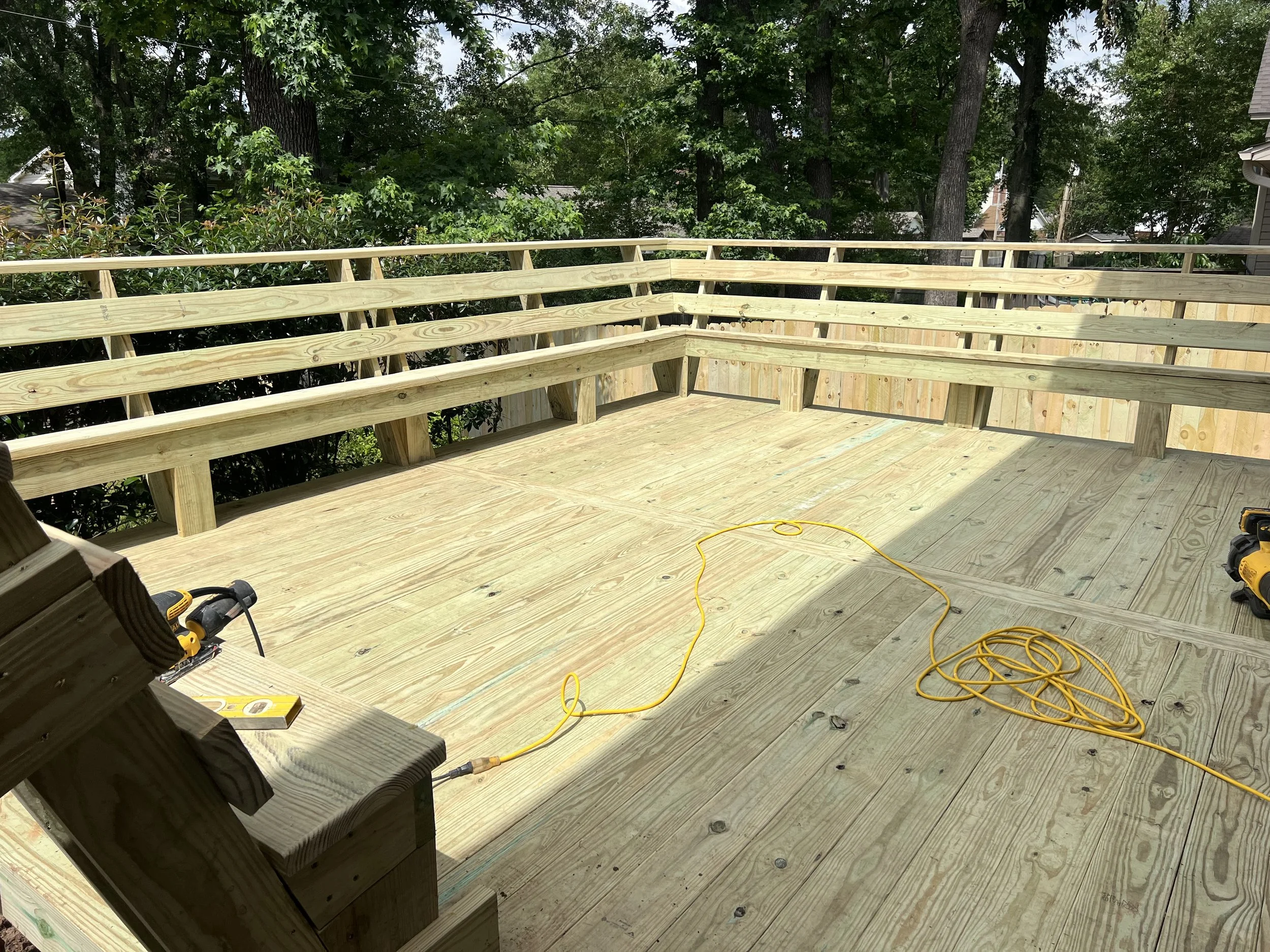 A wooden deck under construction with unfinished railing, construction tools, and extension cords visible, surrounded by trees and residential houses.