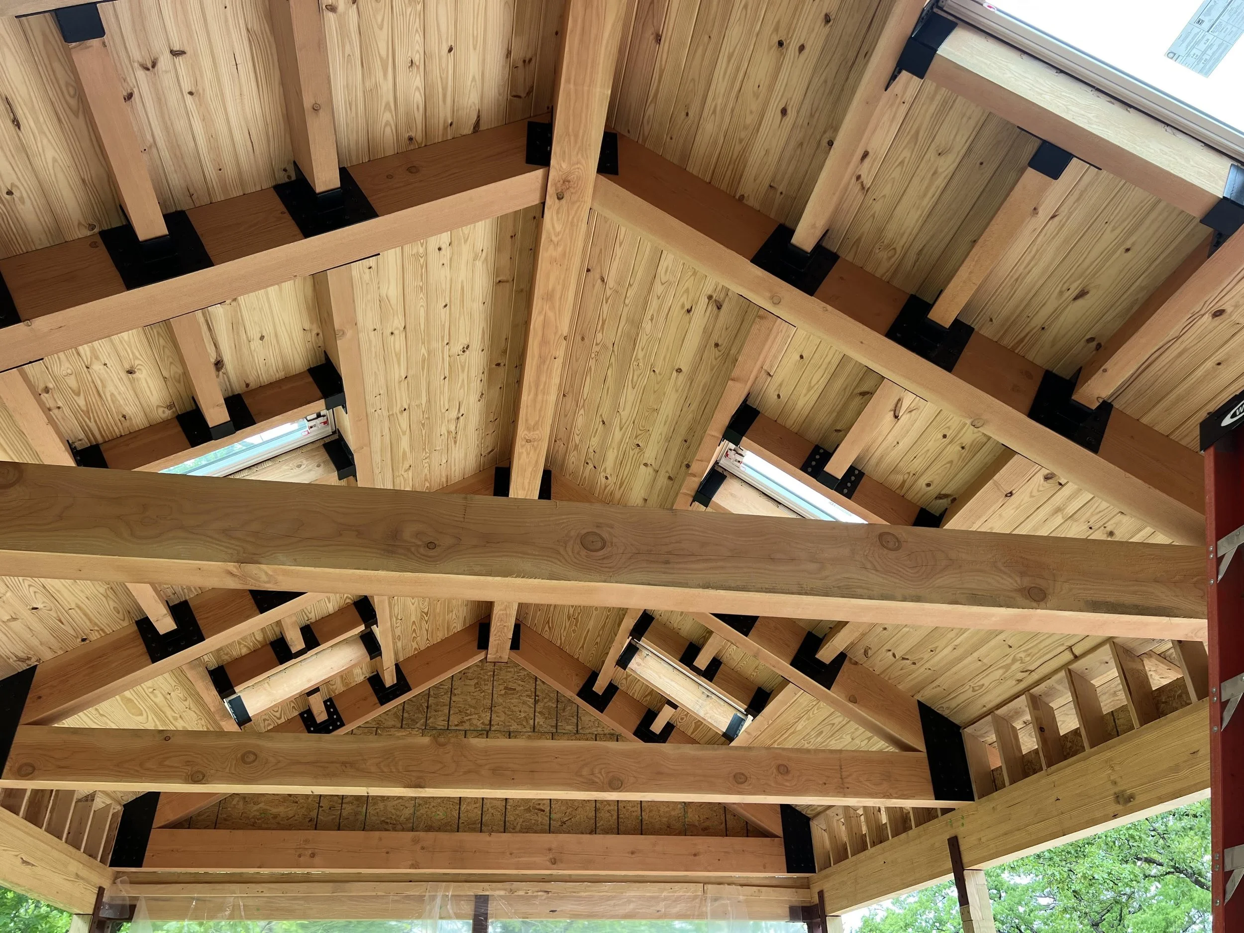Looking up at the underside of a multi-level wooden deck or porch with support beams, joists, and a window at the top right