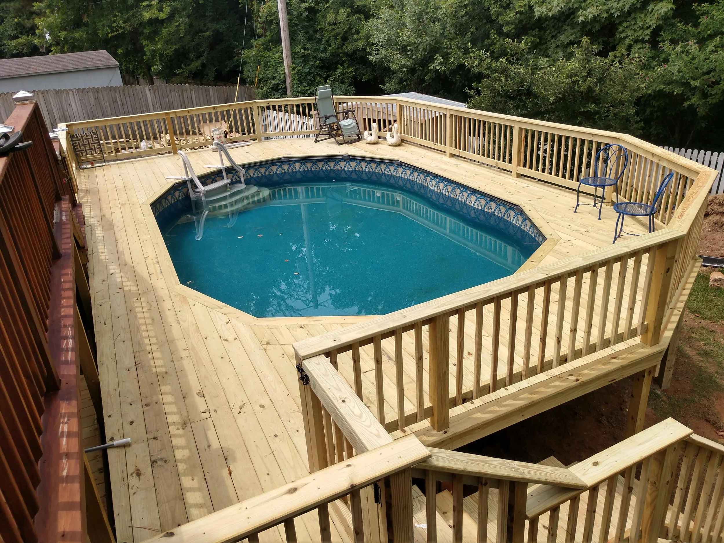 A backyard with a newly built wooden deck around an above-ground swimming pool. The deck features stairs leading down and contains outdoor furniture and garden decorations, with trees and a fence in the background.