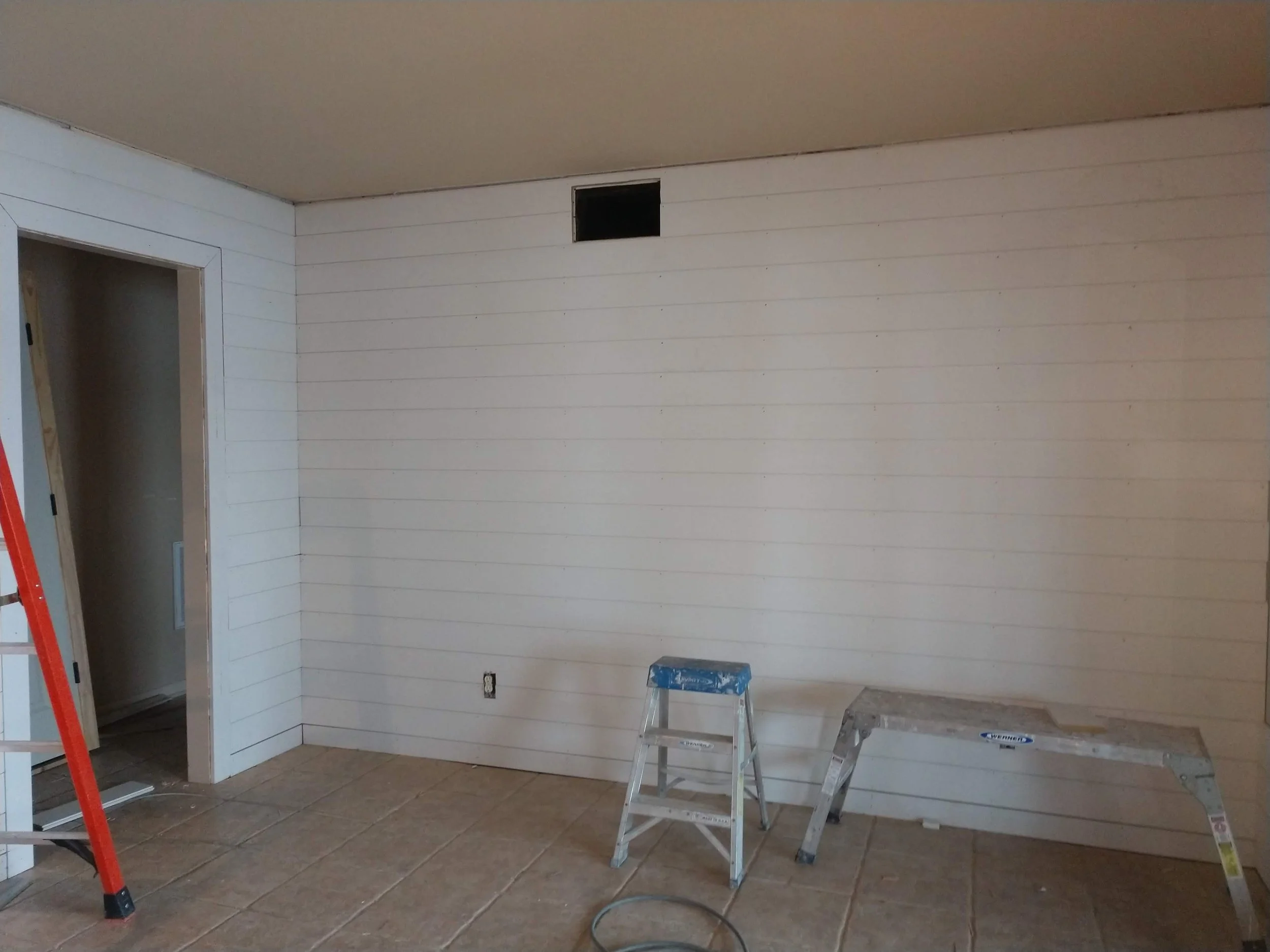 Interior room under renovation with white shiplap walls, a small square vent near the ceiling, a partially open door frame, and construction tools including a step ladder, a small scaffold, and a red broom.